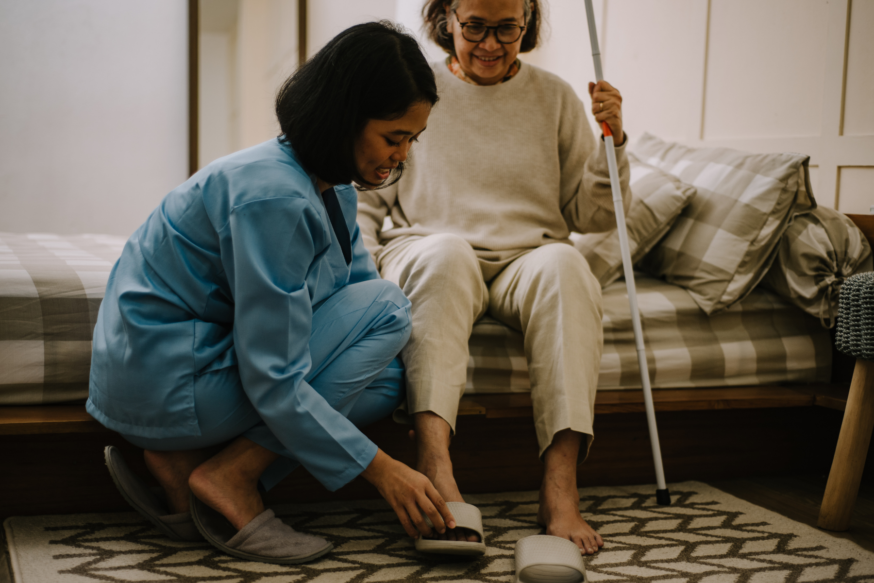 A young woman in blue pajamas helping an elderly woman in beige pajamas put on her slippers on a rug next to a bed.