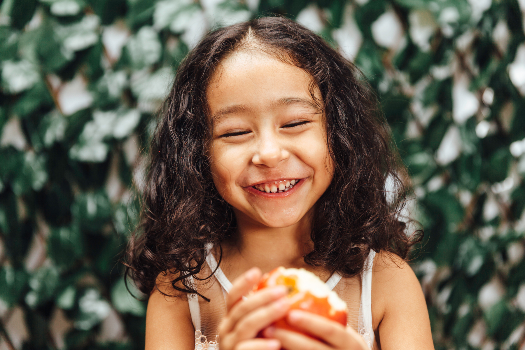 A young girl with curly dark hair smiling while holding a piece of food, standing in front of a leafy green background.