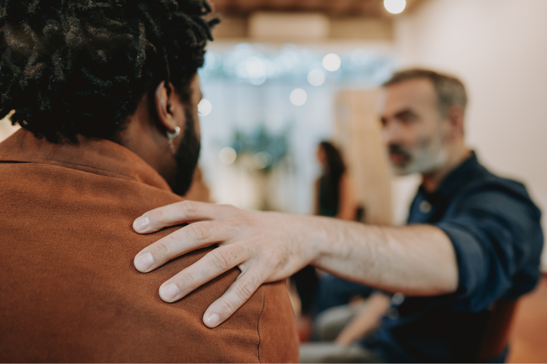 A person with dark skin and curly hair receiving a handshake from a man with gray hair and a beard in a blurred indoor setting.