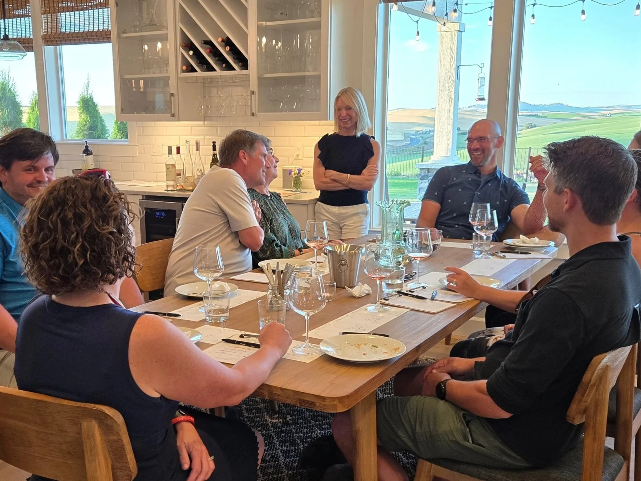 People gathered around a dining table in a bright room, listening to a woman speaking, with wine glasses and table settings, in front of large windows showing a scenic outdoor view.