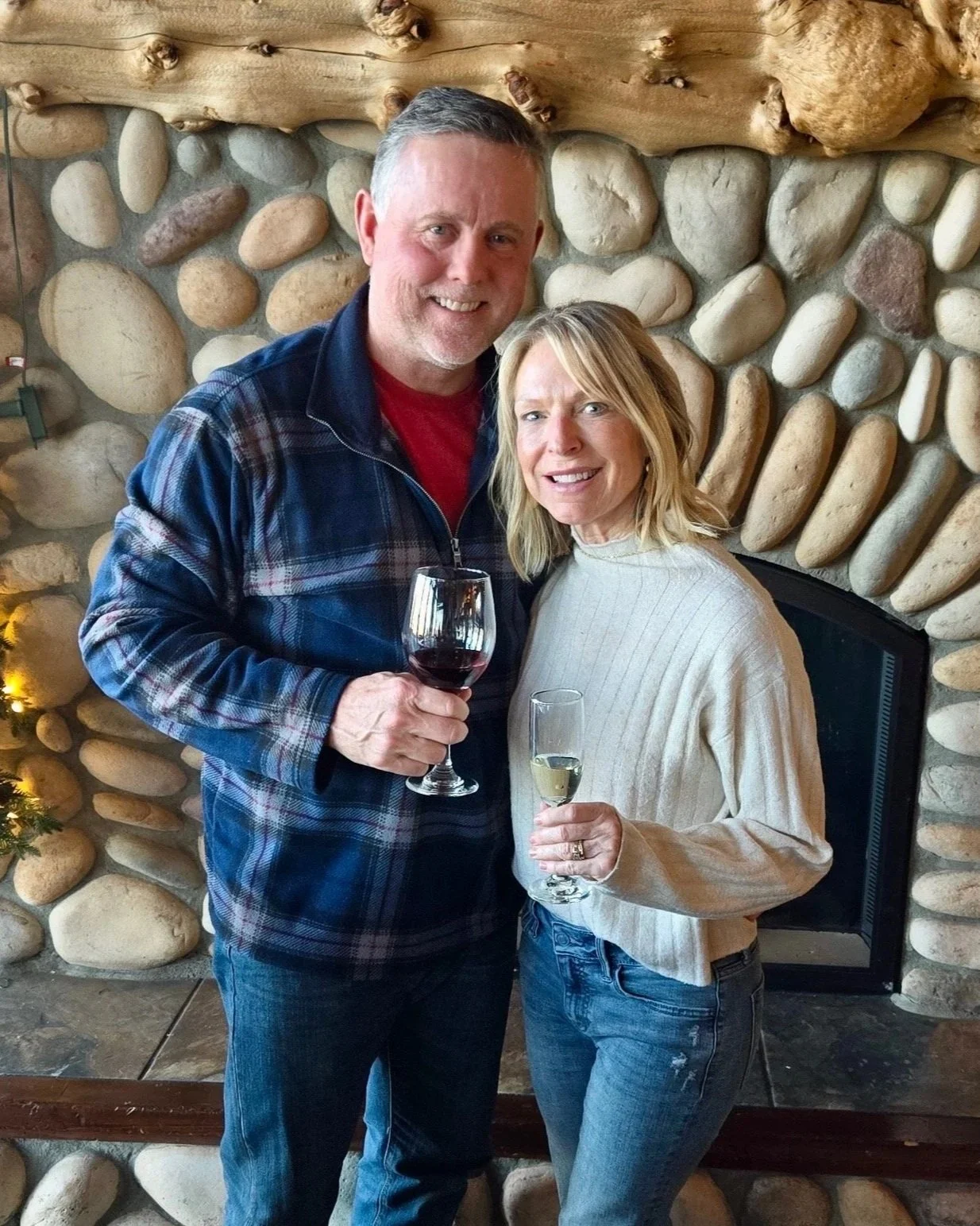 A smiling Bob and Sheryl Bashaw holding glasses of wine and champagne, standing in front of a stone fireplace with a wooden mantle.