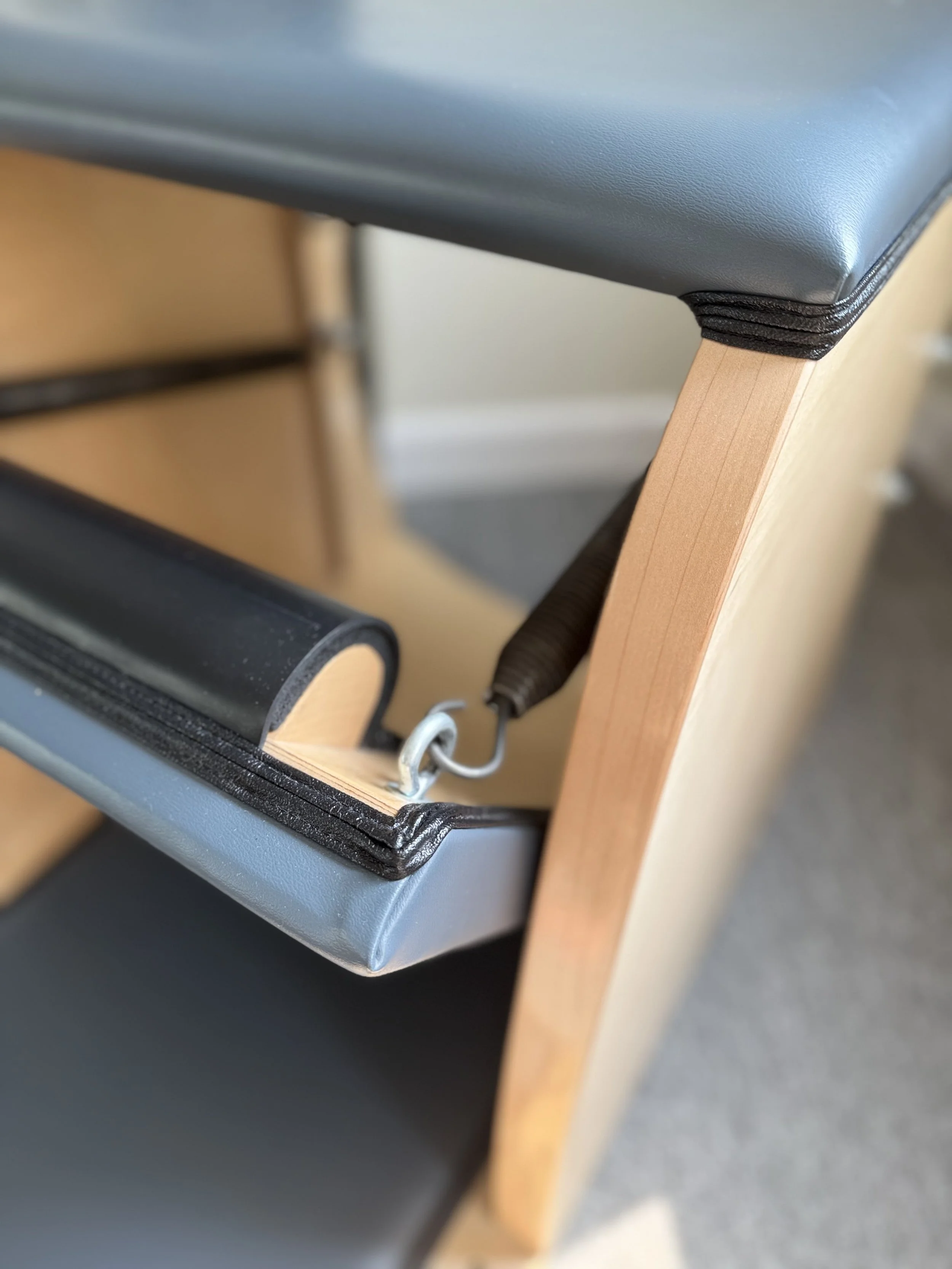 Close-up of a wooden exercise bench with a black padded surface and a black strap, showing part of the frame and hinge mechanism.