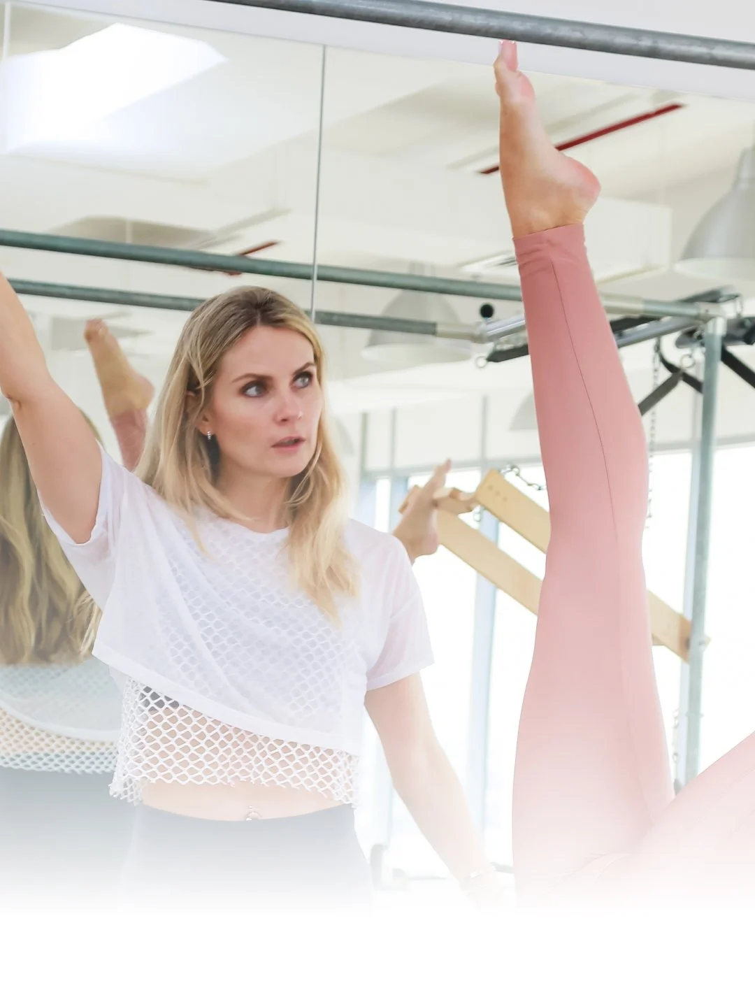 A woman with blonde hair observing or instructing a person doing a leg stretch in a fitness or yoga class.