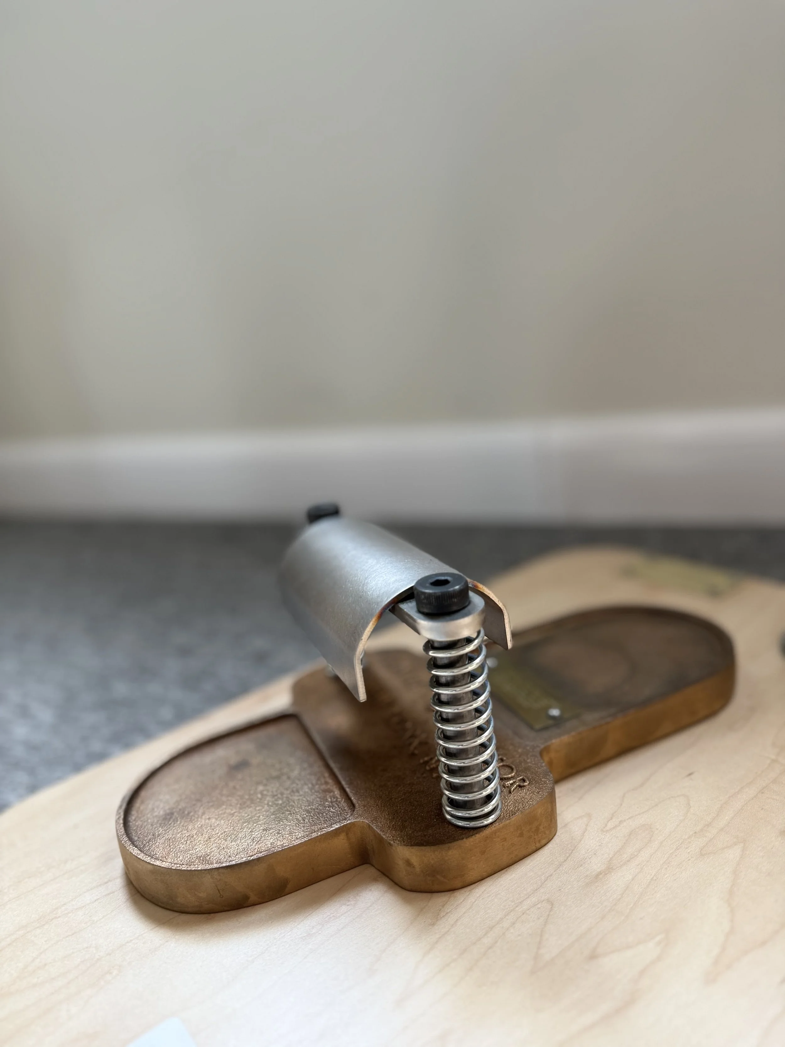 Close-up of a metallic spring attached to a wooden base, with a metal fixture on top, placed on a gray surface with a plain beige wall in the background.