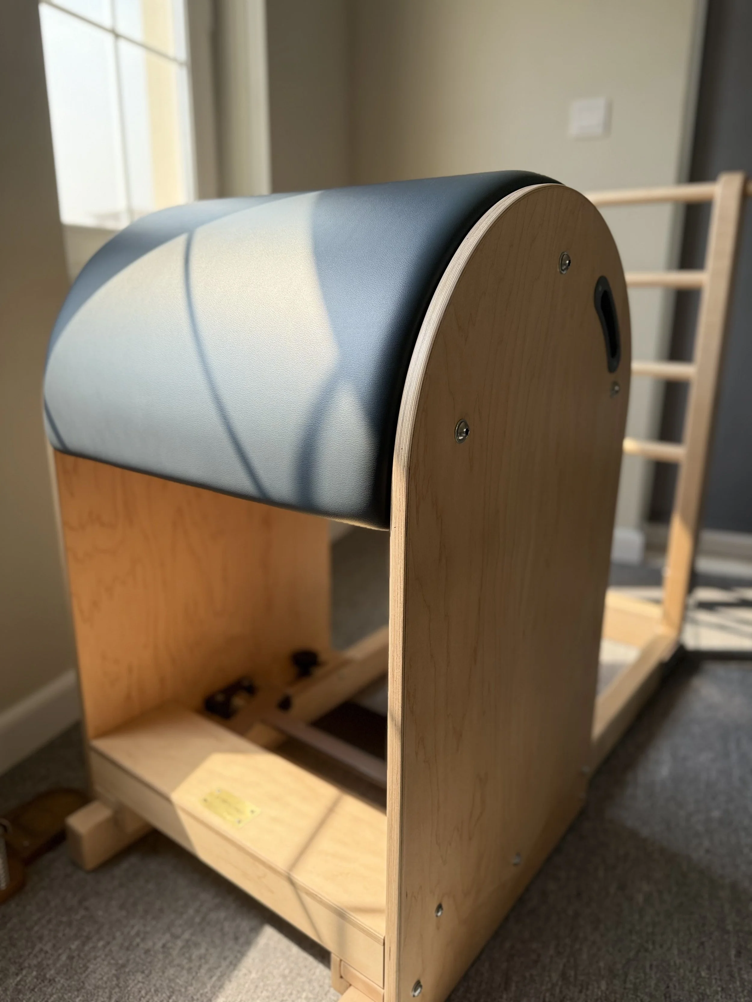 An incomplete wooden arcade-style game cabinet with a curved top, missing its screen and other components, placed on a carpeted floor near a window and a wall.