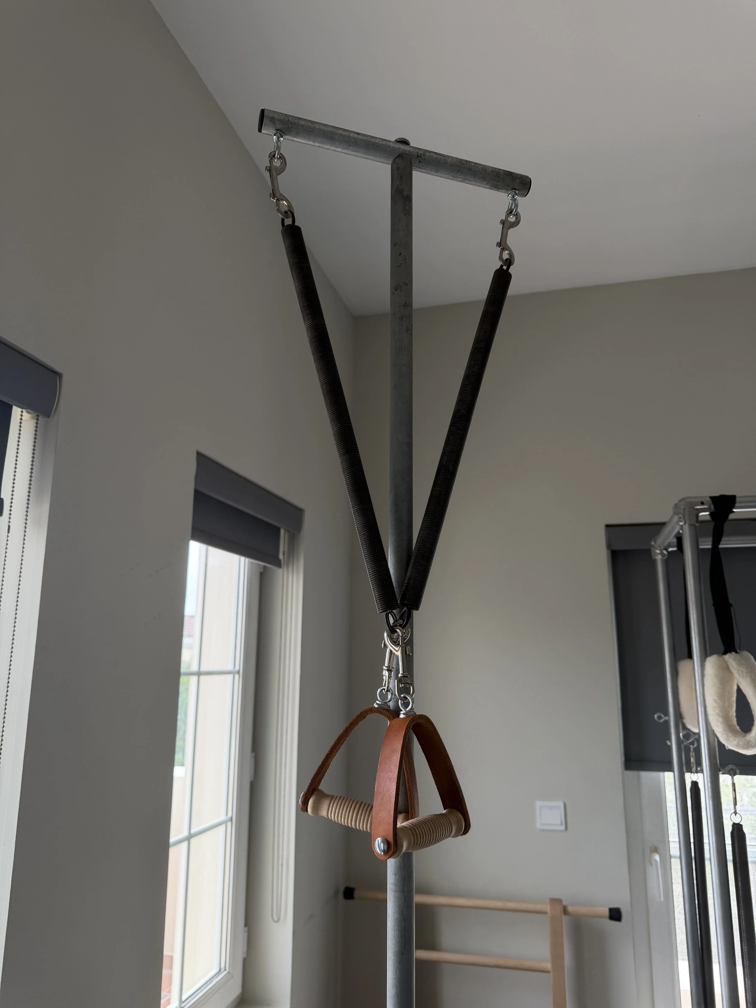 Gym exercise equipment with suspension straps and handles hanging from a ceiling mount in a workout room with large windows.