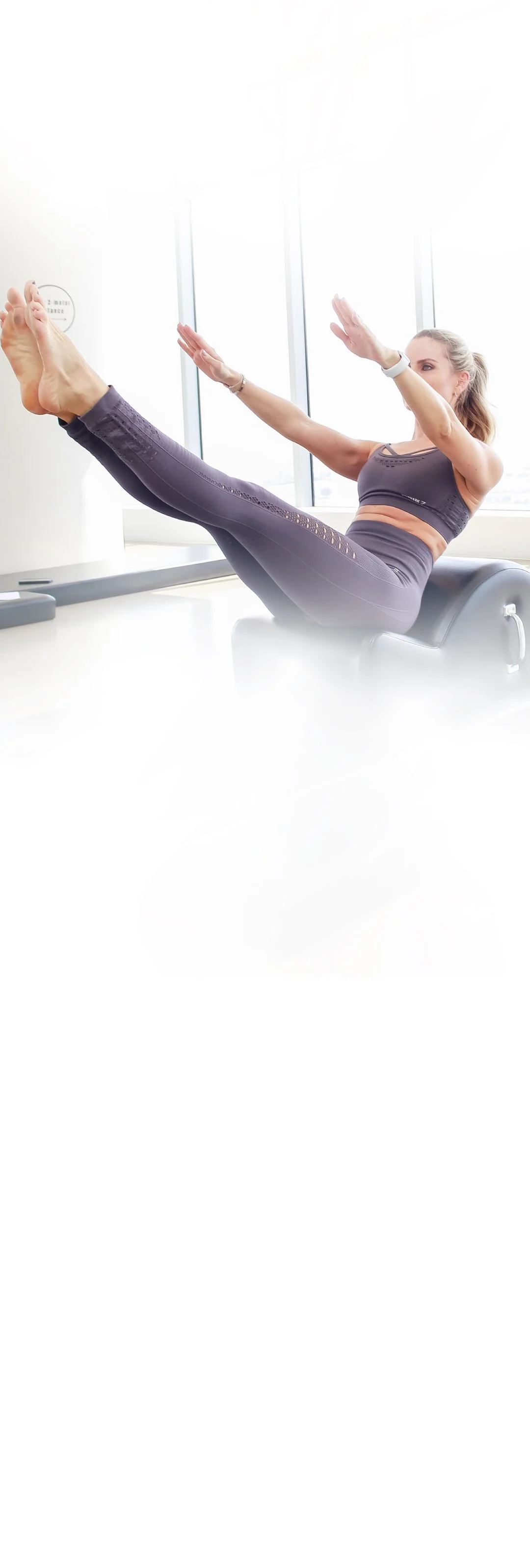 Woman in workout attire doing Pilates or core exercise on equipment near large windows.