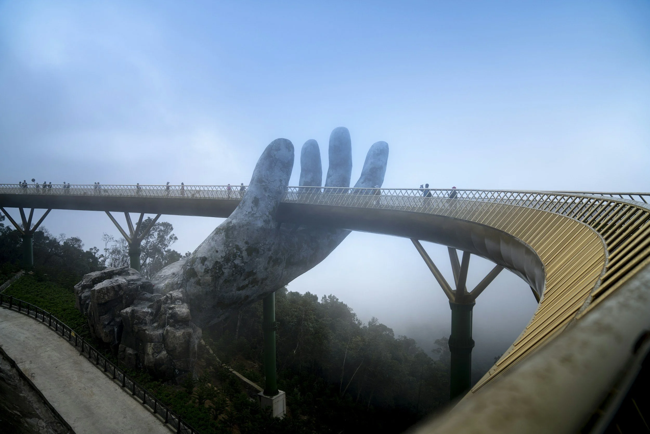A large pedestrian bridge supported by green pillars, designed to look like a giant open hand emerging from a rocky landscape, with a misty sky in the background.