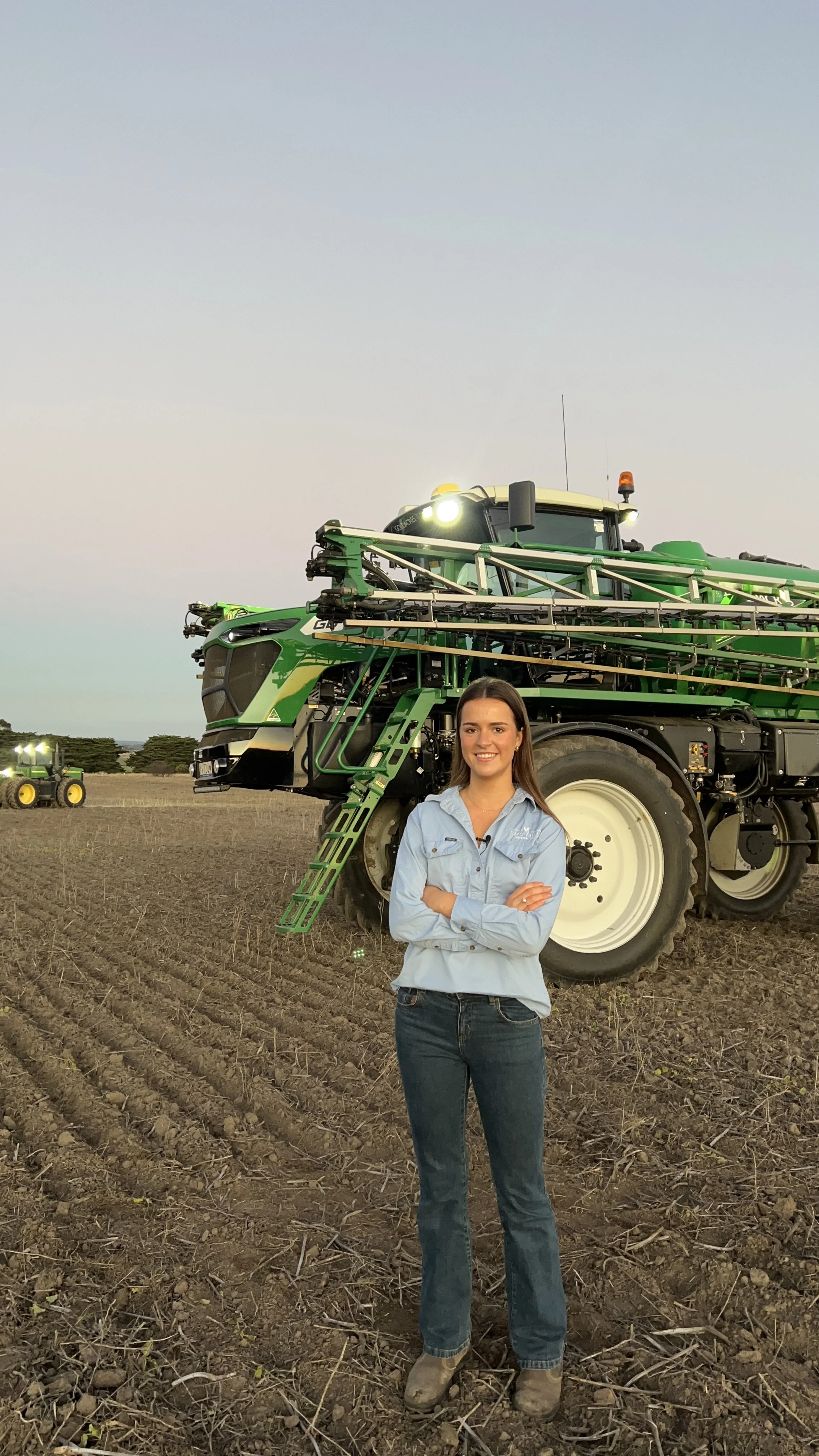 A woman standing in a field with arms crossed in front of a large green tractor, during sunset with clear sky and other tractors in the background.