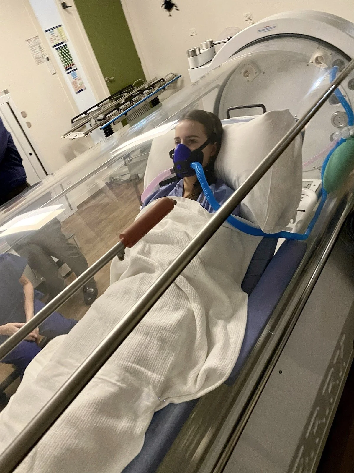 A woman lying in a medical incubator with a breathing mask connected to a ventilator, in a hospital room.