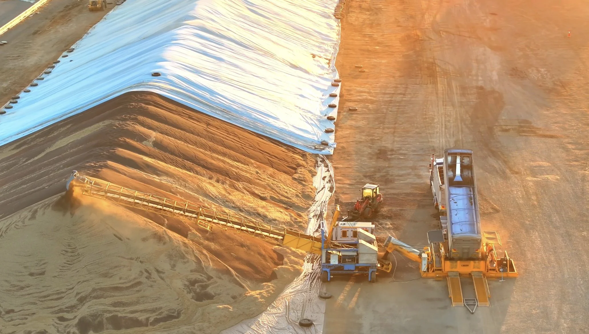Large industrial machine spreading sand or soil on a construction or excavation site with vehicles nearby, under warm sunset light.