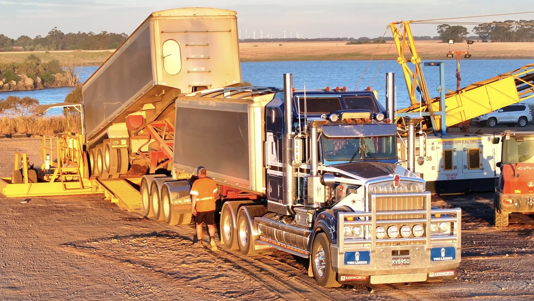 A large black semi-truck with a chrome grille and multiple lights on the front is parked on dirt near a body of water. The truck is connected to a yellow conveyor system used for loading or unloading, and a person in an orange safety vest and black shorts stands nearby. There are other construction vehicles and equipment, with a rural landscape and lake in the background.