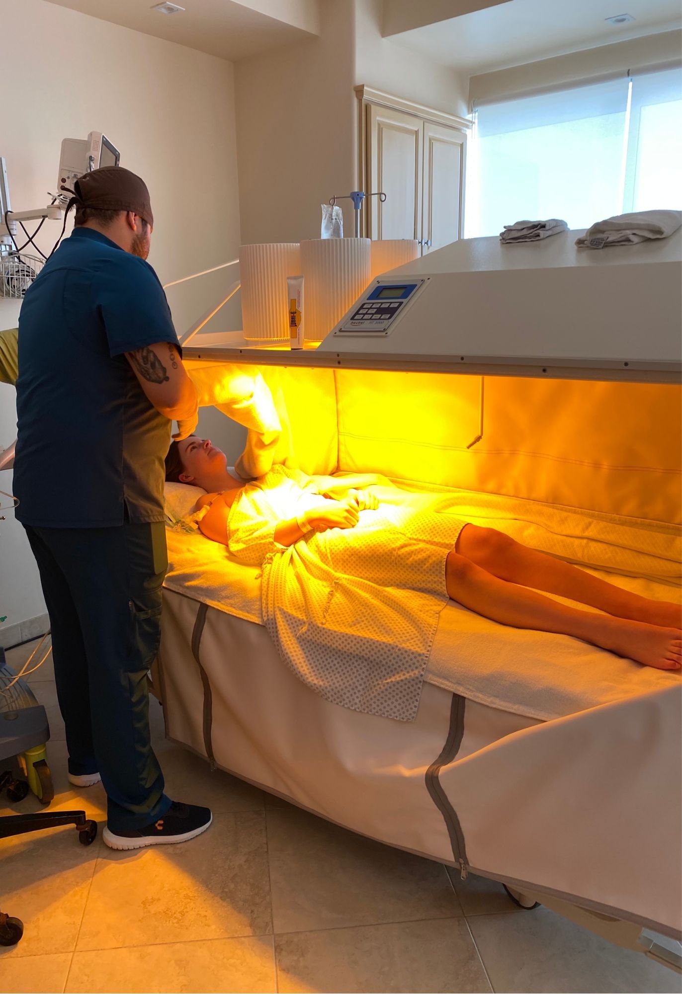 A person lying on a bed inside a hyperbaric oxygen therapy chamber with a medical professional attending to them, under a bright orange light.