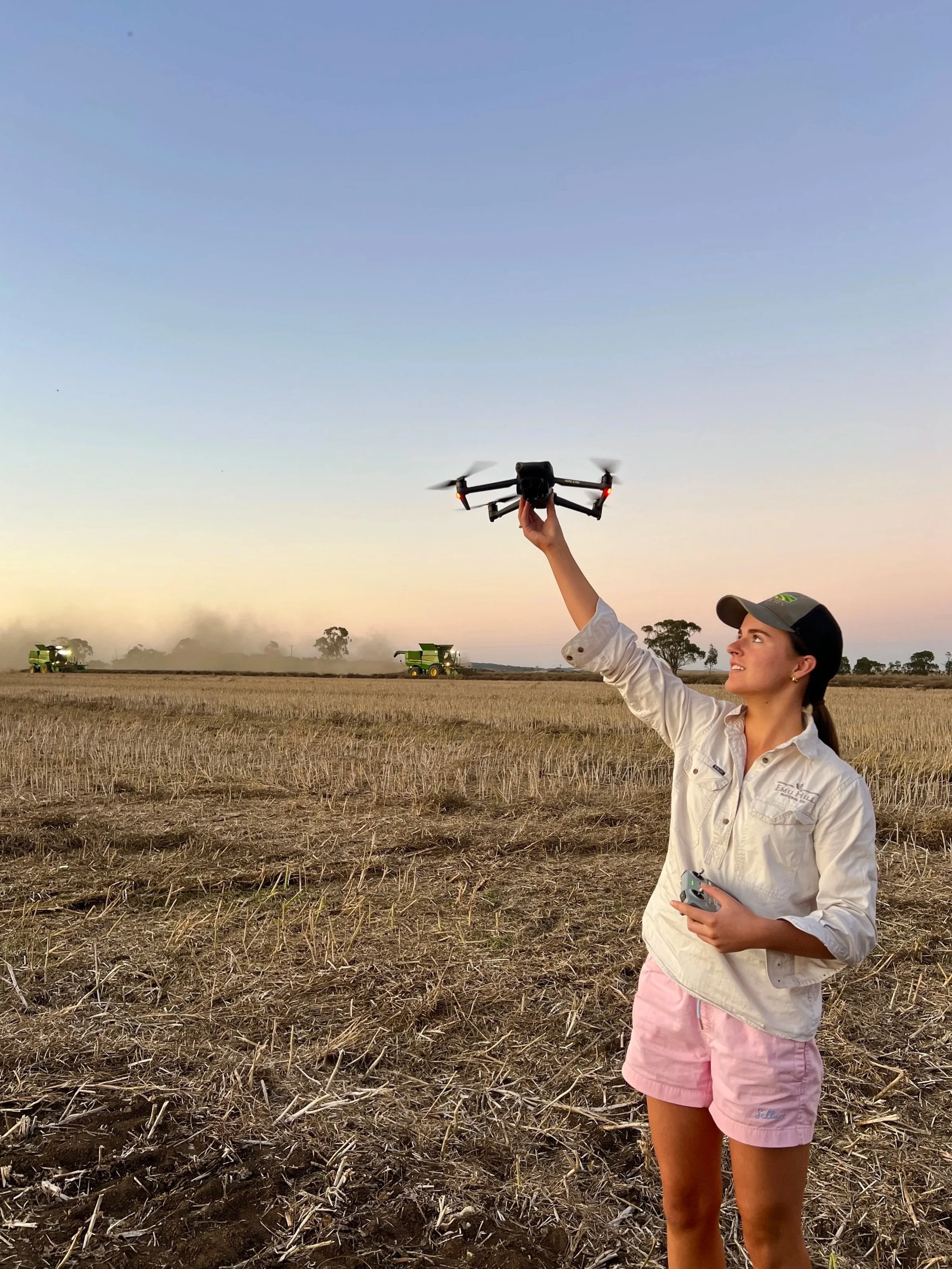 A woman in a light-colored jacket, pink shorts, and a hat is standing in an open field at sunset, launching a drone into the sky.