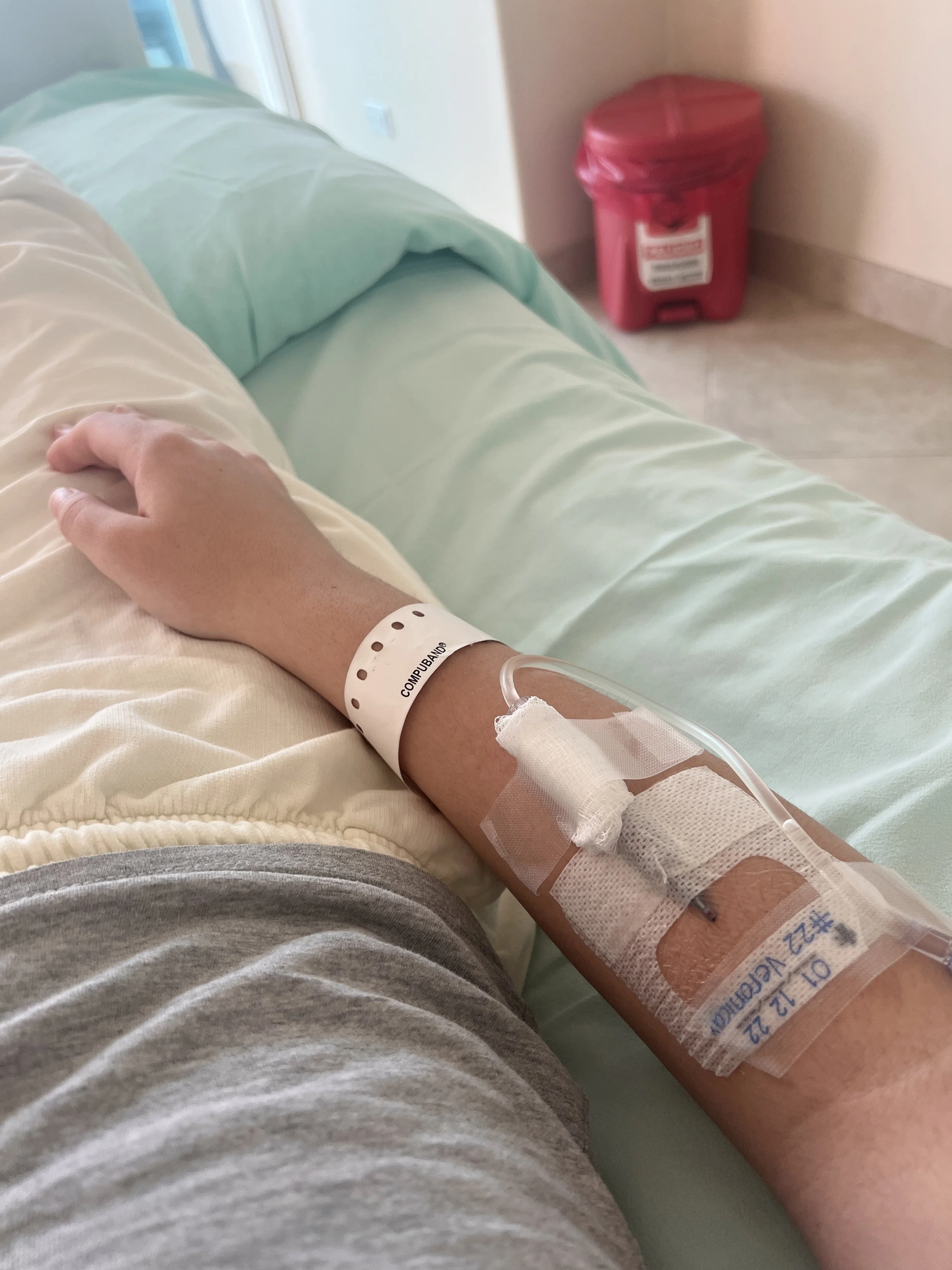 A hospital patient's arm with an IV inserted, lying on a hospital bed with light green sheets. A white wristband is on the wrist and a red sharps disposal container is visible in the background in the hospital room.