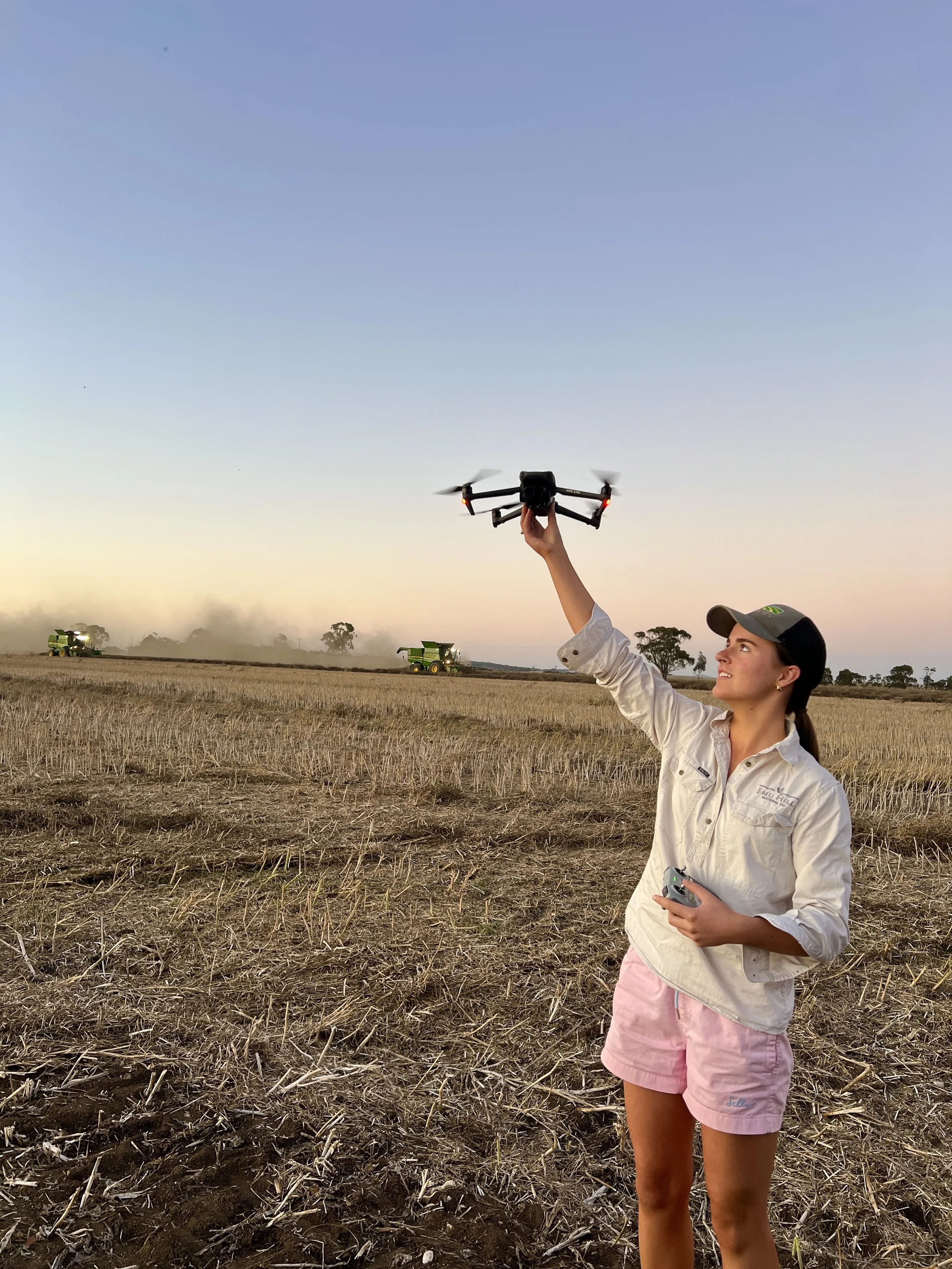 Young woman in farm attire flying a drone in a harvested field during sunset with farming machinery in the background.