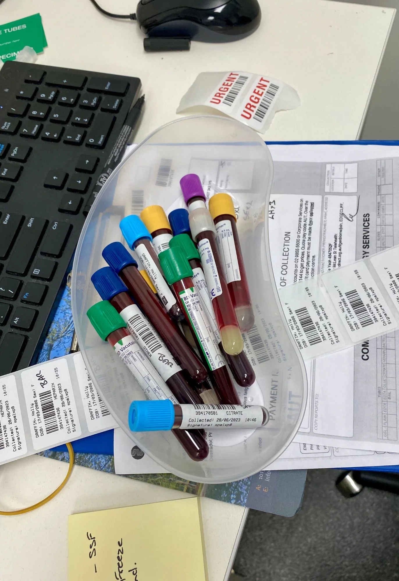 Blood collection tubes with colored caps on a cluttered office desk, alongside medical paperwork, a keyboard, a computer mouse, urgent labels, and a yellow sticky note with handwritten notes.