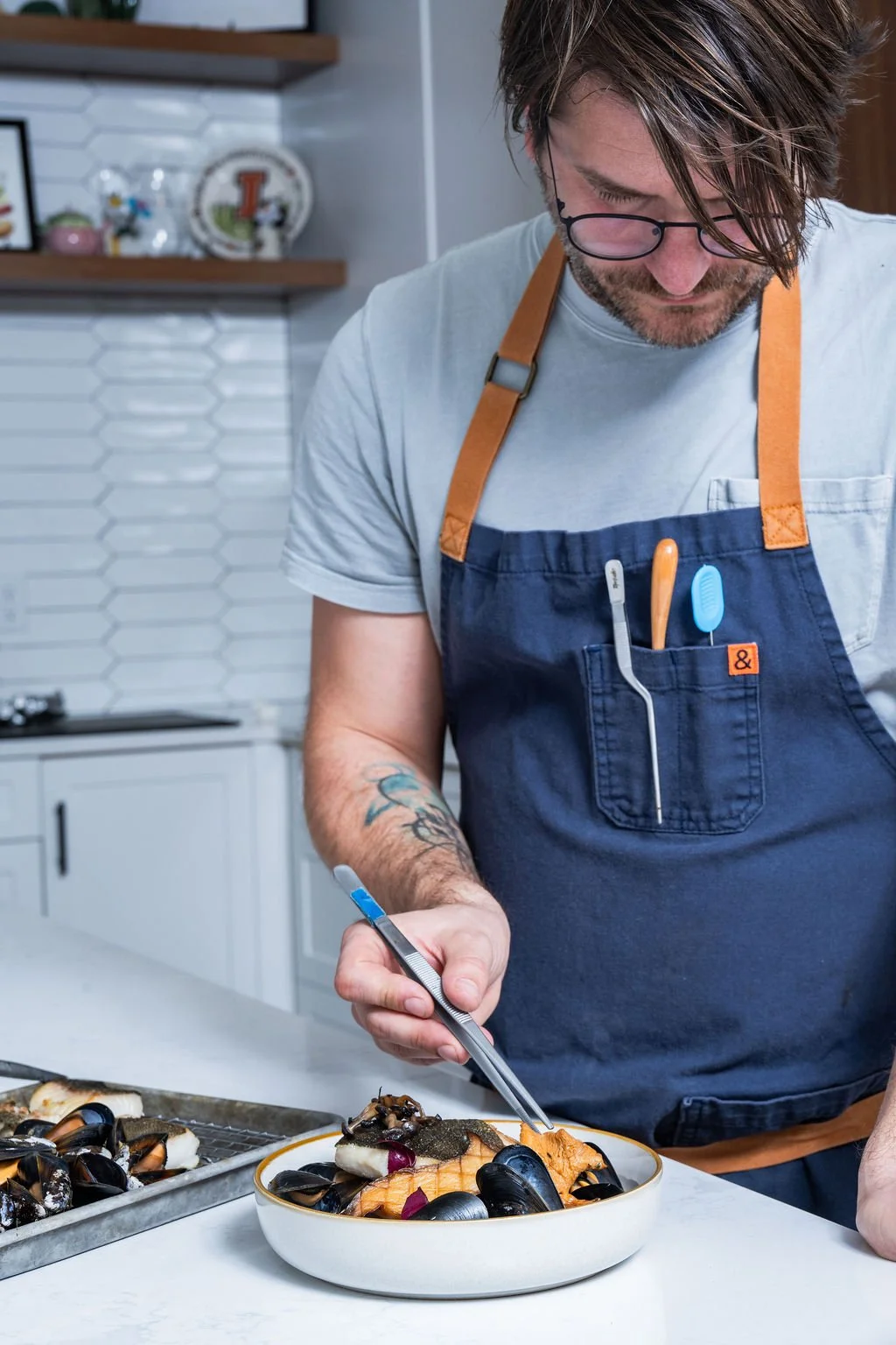 A man with glasses and a tattoo on his left arm is preparing a seafood dish in a kitchen. He is wearing a navy apron with utensils in the pocket, including a toothbrush and a spoon, and using tongs to place seafood on a plate. There are more seafood items on a tray in front of him.