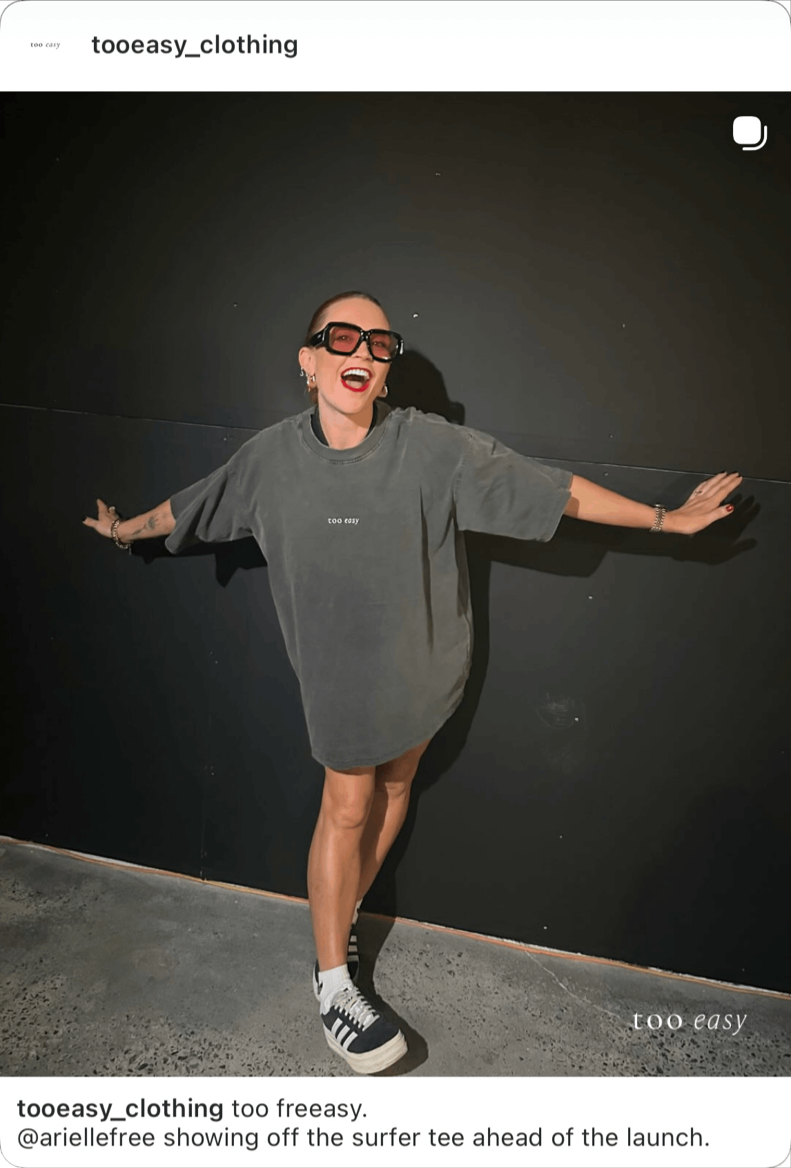 Woman in oversized gray t-shirt with 'too easy' text, shorts, sneakers, large glasses, and jewelry, standing against a black wall with arms outstretched, smiling and showing off her outfit.