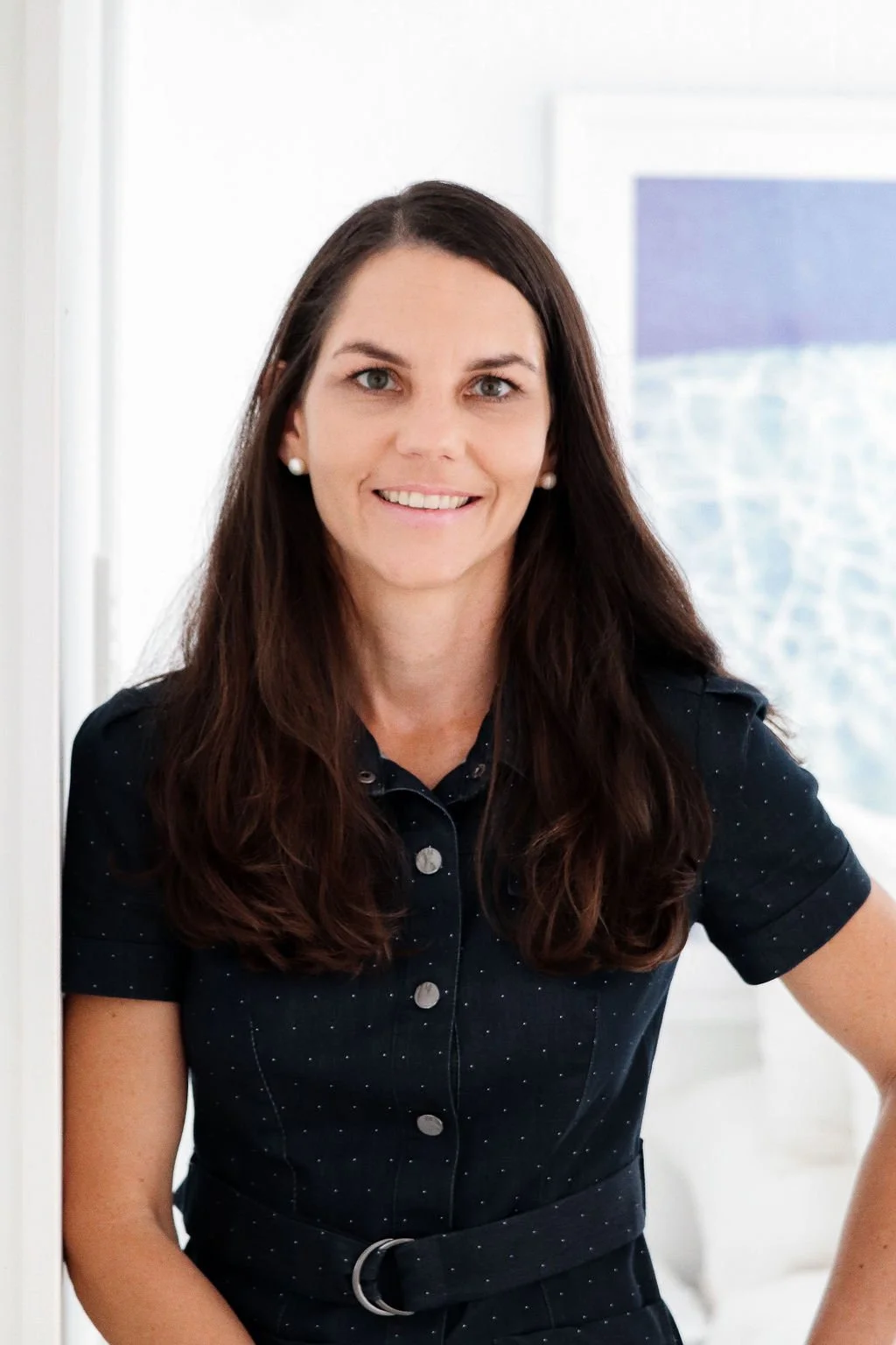 Natalie Kidcaff consulting and coaching with long dark hair and pearl earrings smiling, wearing a black dress with small white polka dots, standing indoors near a window.