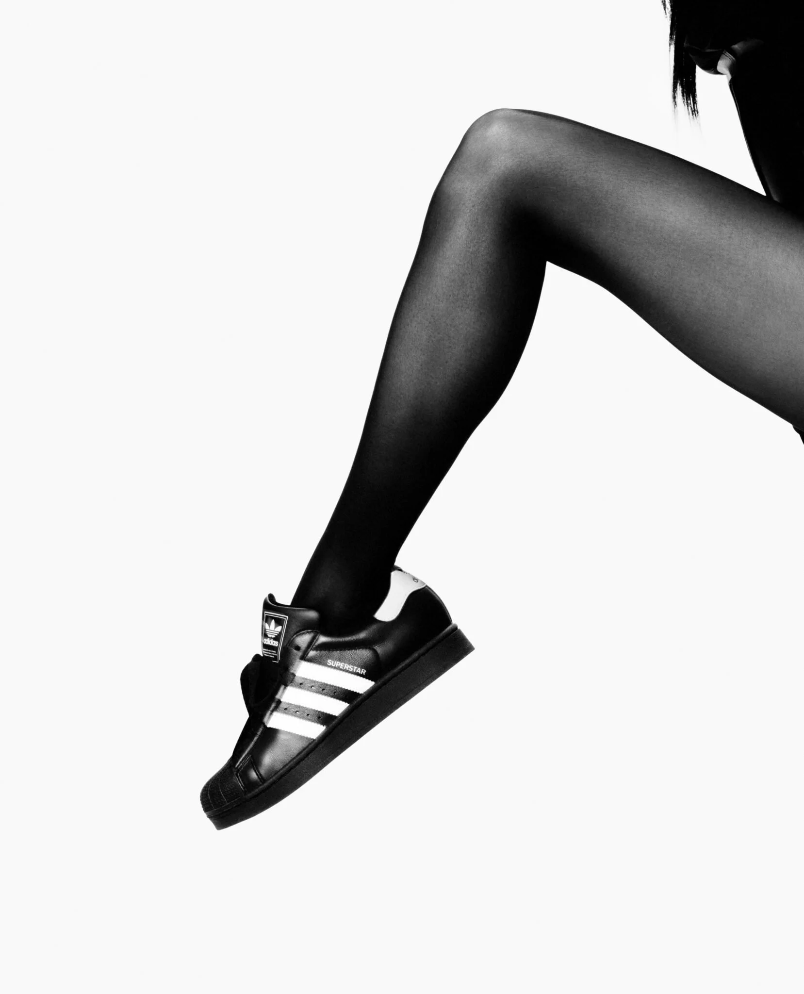 Close-up of a woman’s leg wearing black stockings and an adidas Superstar sneaker, with a plain white background.