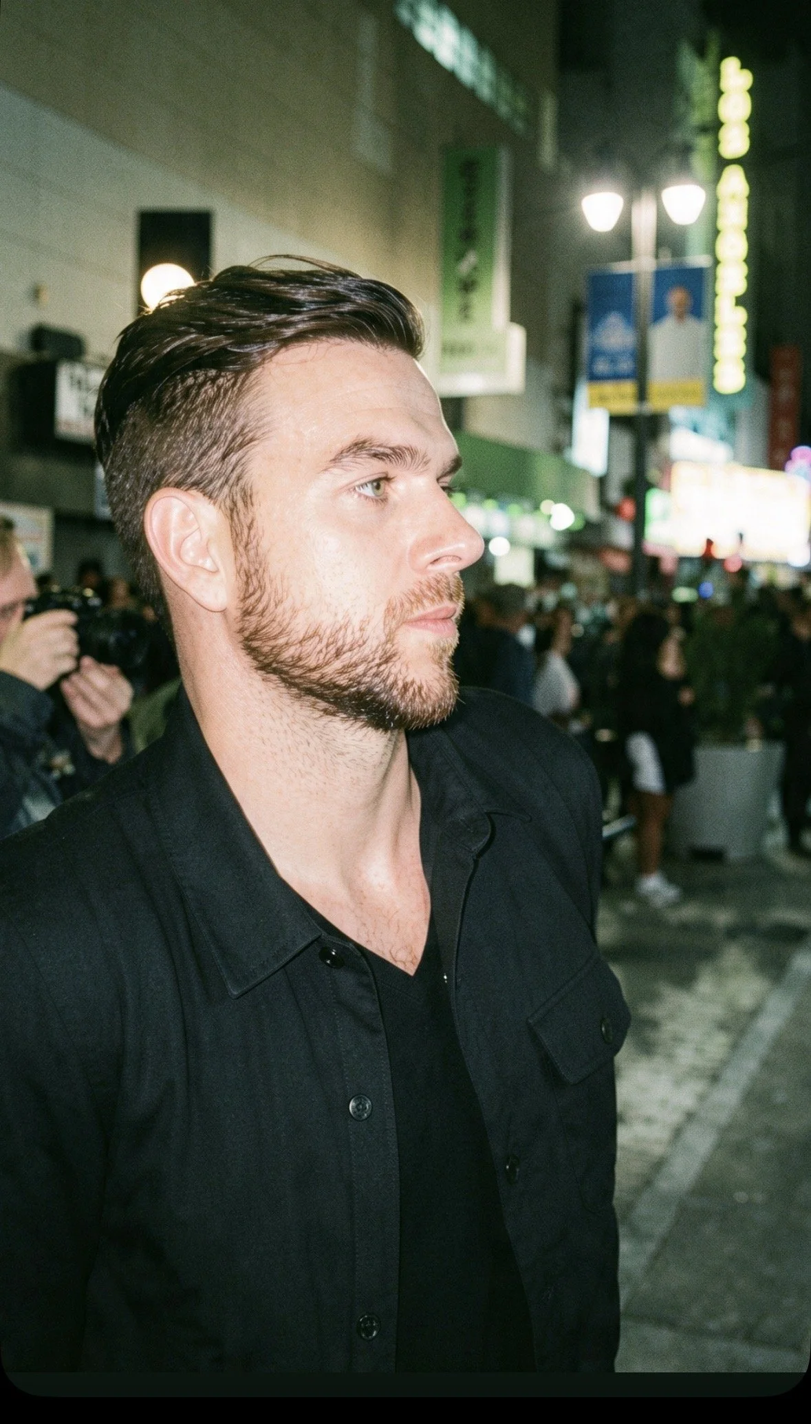 Bearded man in a black jacket photographed in profile at a busy nighttime event.