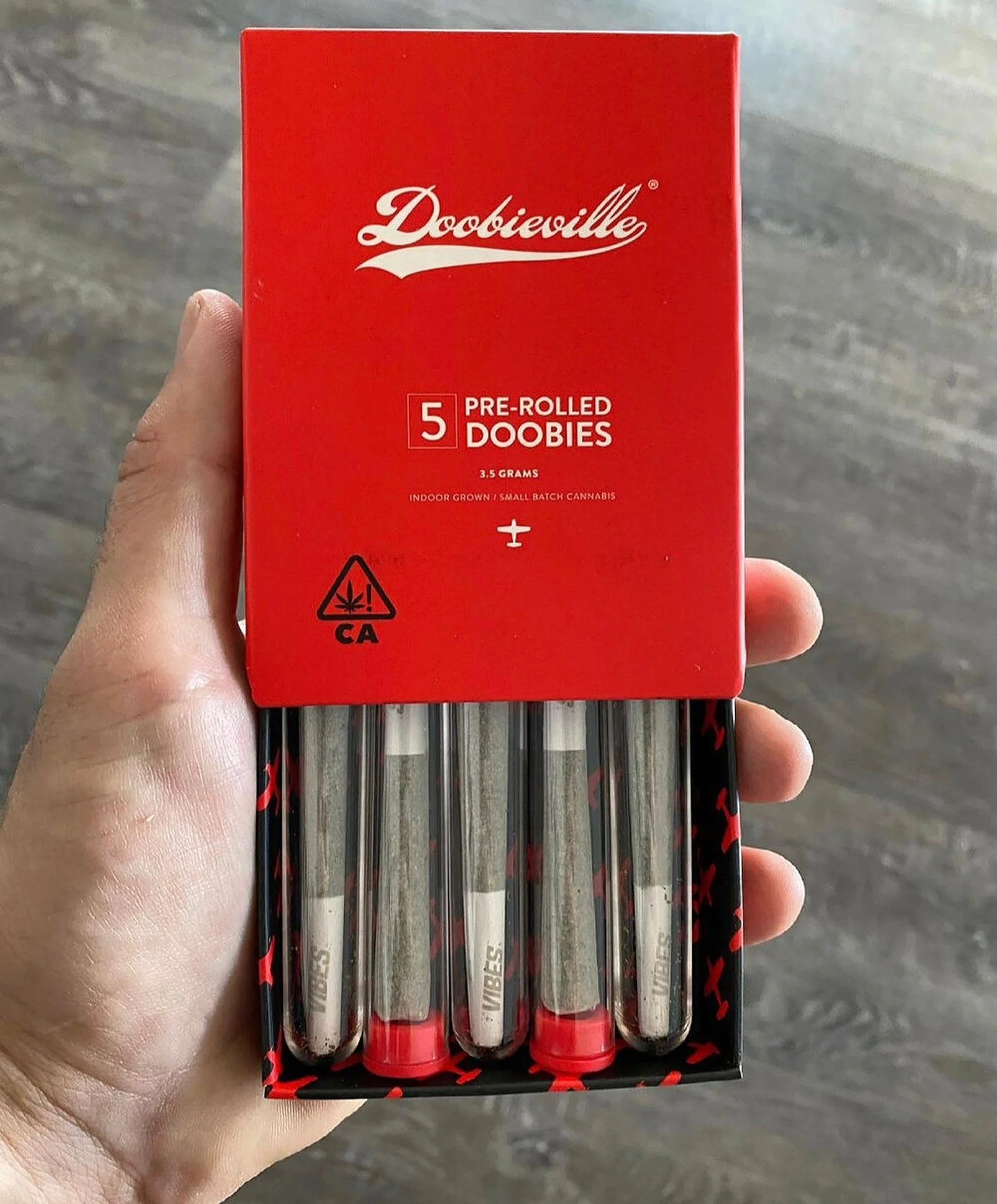 A hand holding a red box of Doobie pre-rolled cannabis joints, containing five joints, with a gray wooden surface in the background.