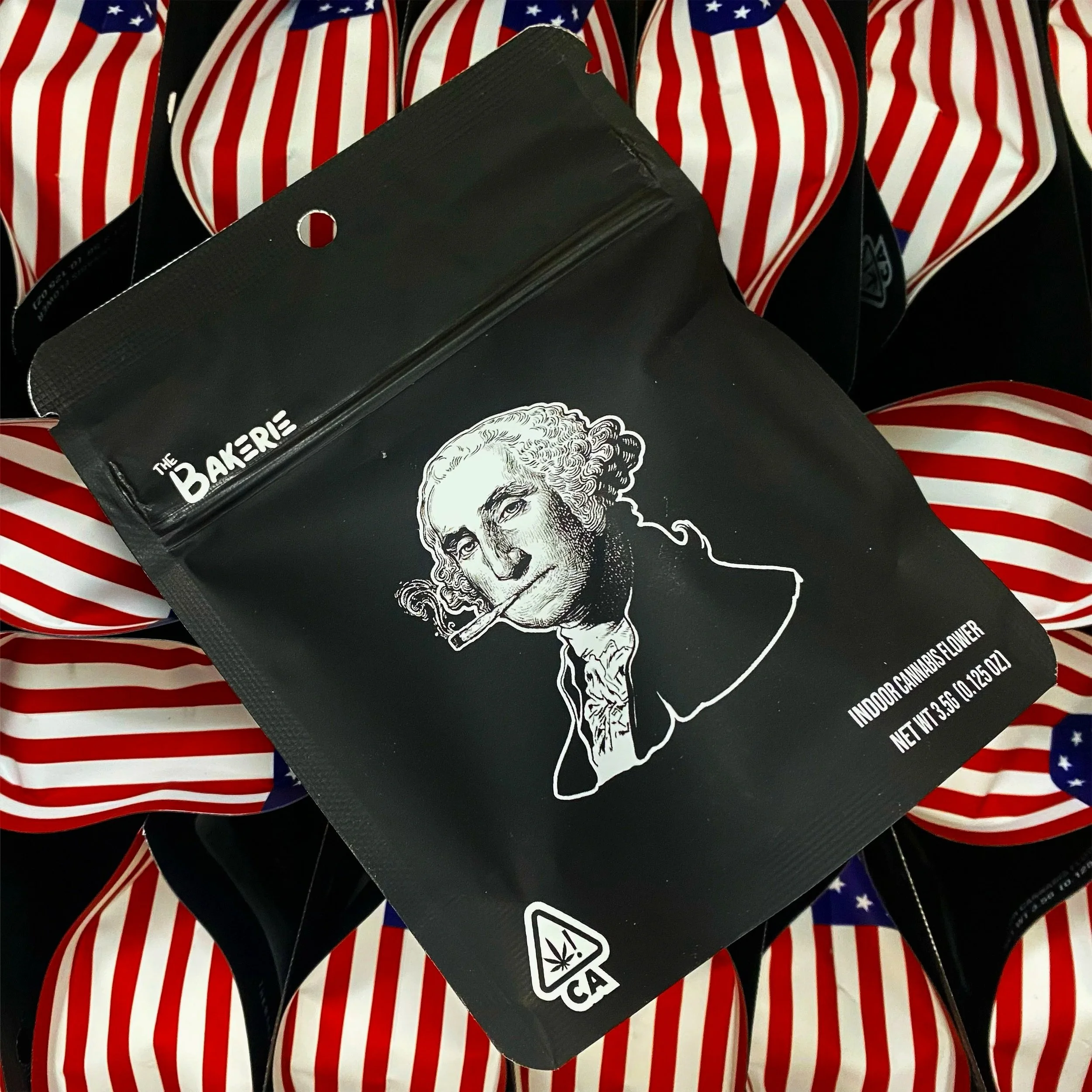 Black package with a stylized drawing of George Washington smoking a joint, labeled 'The Bakerie' and 'Indoor Cannabis Flower,' surrounded by American flags.