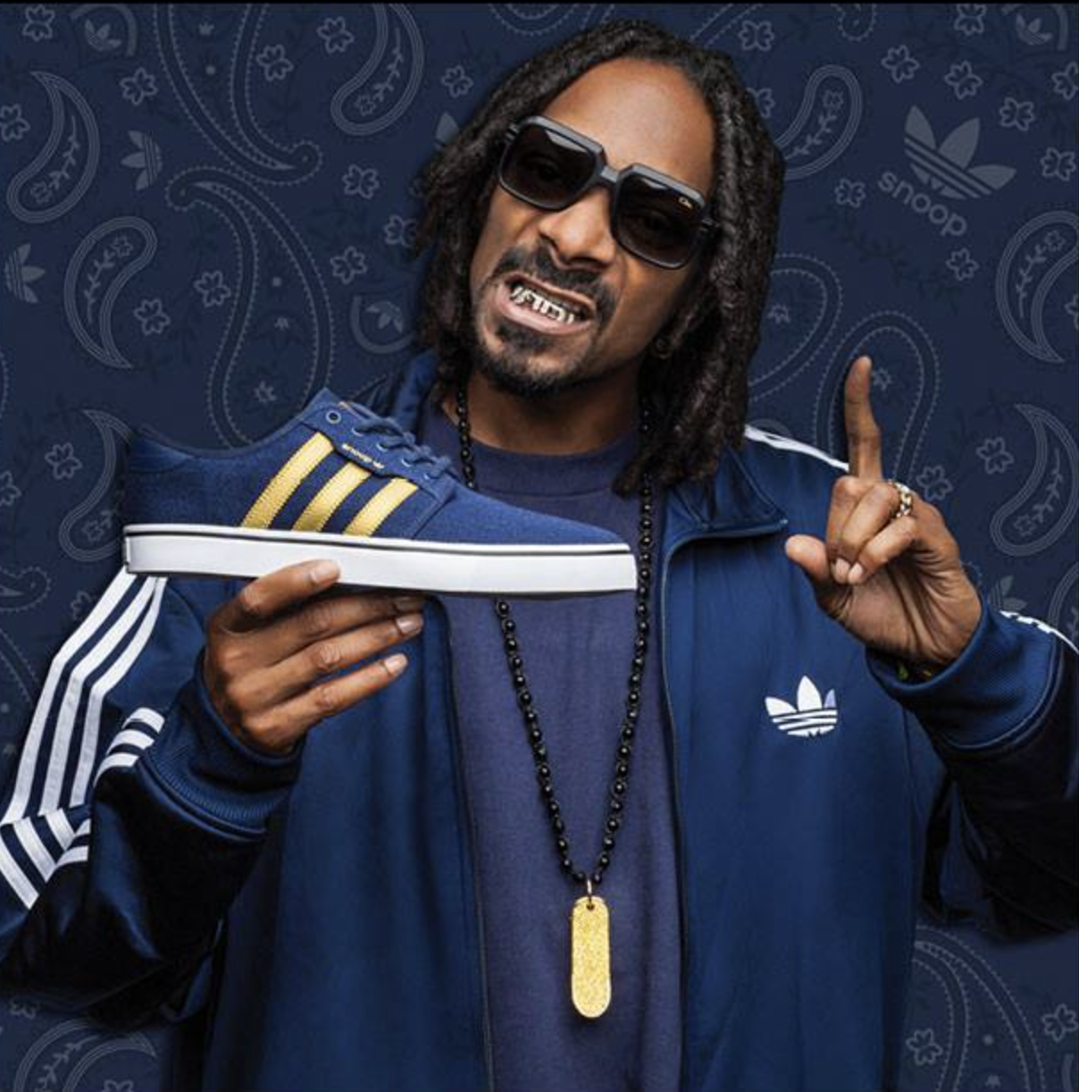 Man wearing sunglasses, a blue Adidas tracksuit, holding a blue sneaker with yellow stripes, pointing upward, against a patterned blue background.