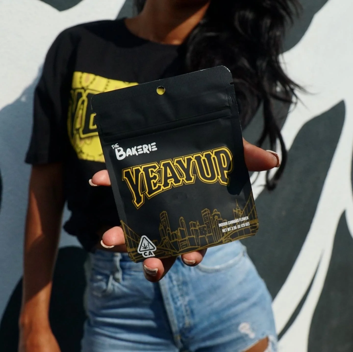 Person holding a black package labeled 'YEAUP' with a cityscape illustration, standing in front of a black and white wall with a design.