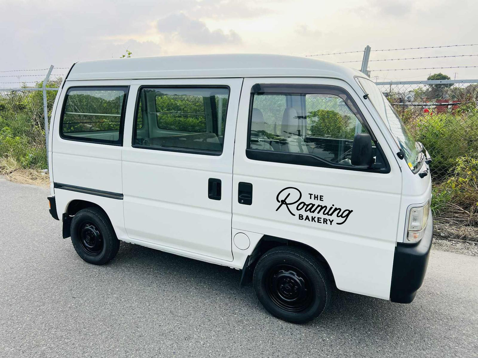 White van with the logo 'The Roaming Bakery' on the side parked on a roadside near a chain-link fence and greenery.