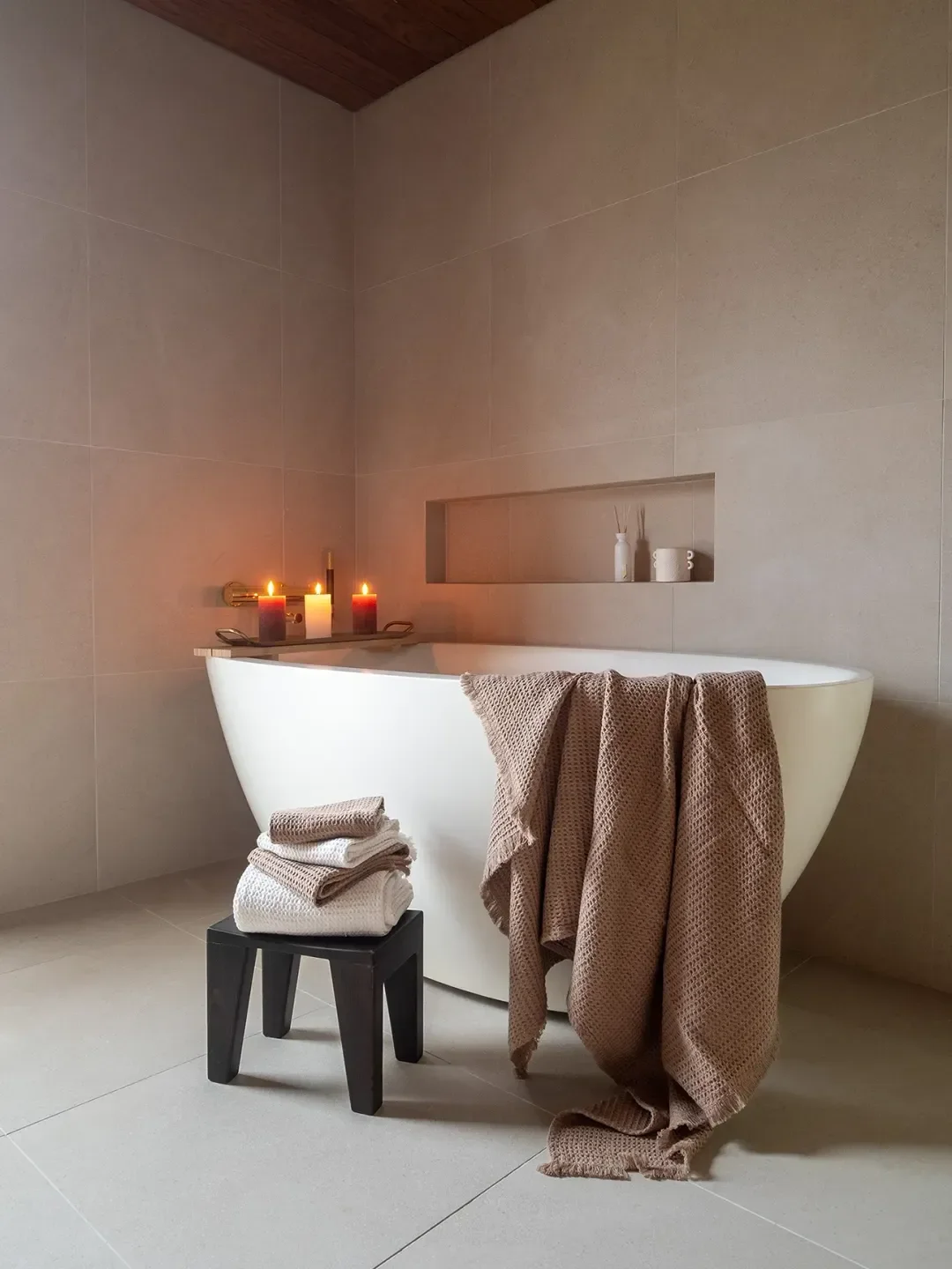 Kuva kylpyhuoneesta, jossa on valkoinen kylpyamme, kynttilöitä, pyyhkeitä pöydällä, ja sisustuselementtejä hyllykössä muurilla.