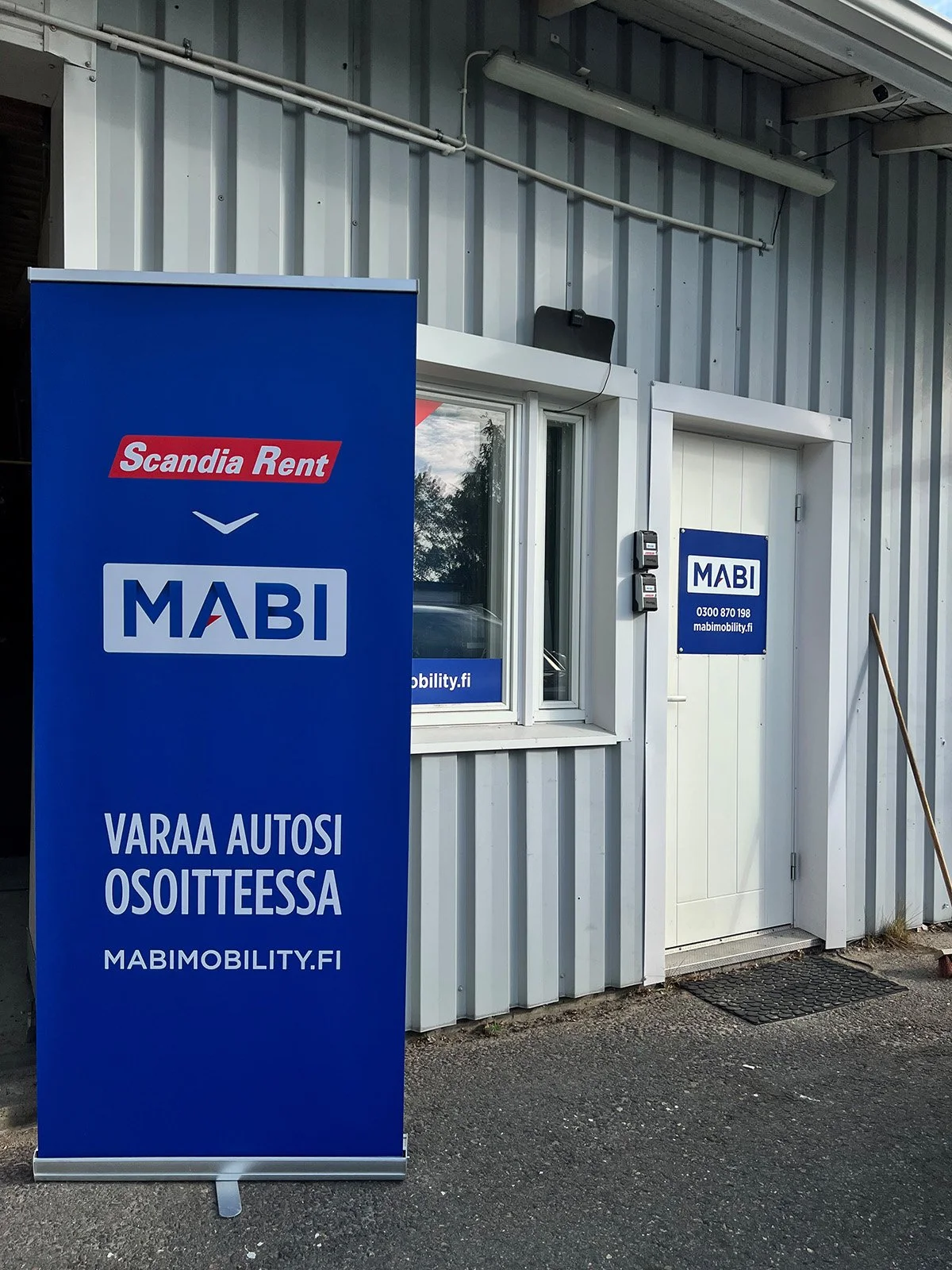Sininen esite, jossa lukee 'Varaa autosi osoitteessa MABIMOBILITY.FI' ja 'Scandia Rent', sijaitsee ulkona pienen rakennuksen edessä.