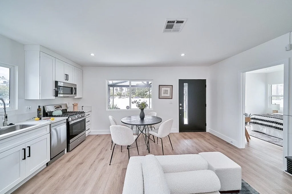 Open concept living room and kitchen with white walls, hardwood floors, dining table with four white chairs, kitchen with stainless steel appliances, and a cozy sitting area.