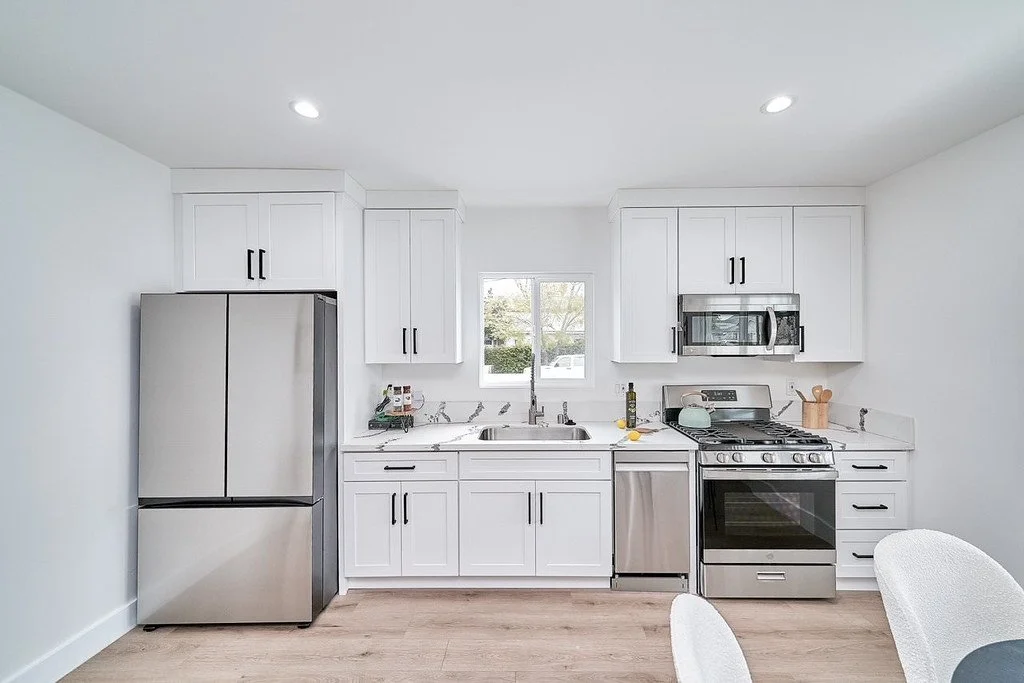 Modern kitchen with white cabinets, stainless steel refrigerator, stove, microwave, and dishwasher. There is a window above the sink, and a small dining table with white chairs in the foreground.