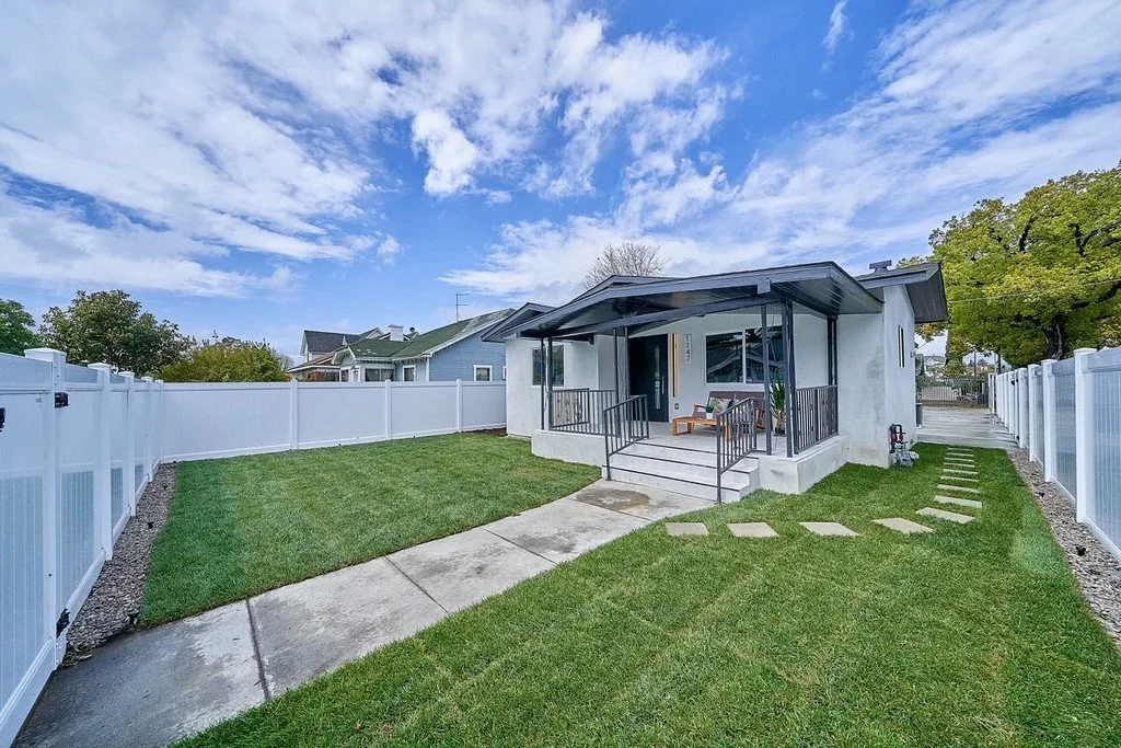 A small house with a porch and a backyard, enclosed by a white fence, with a pathway leading to the porch, green grass, and a partly cloudy sky.