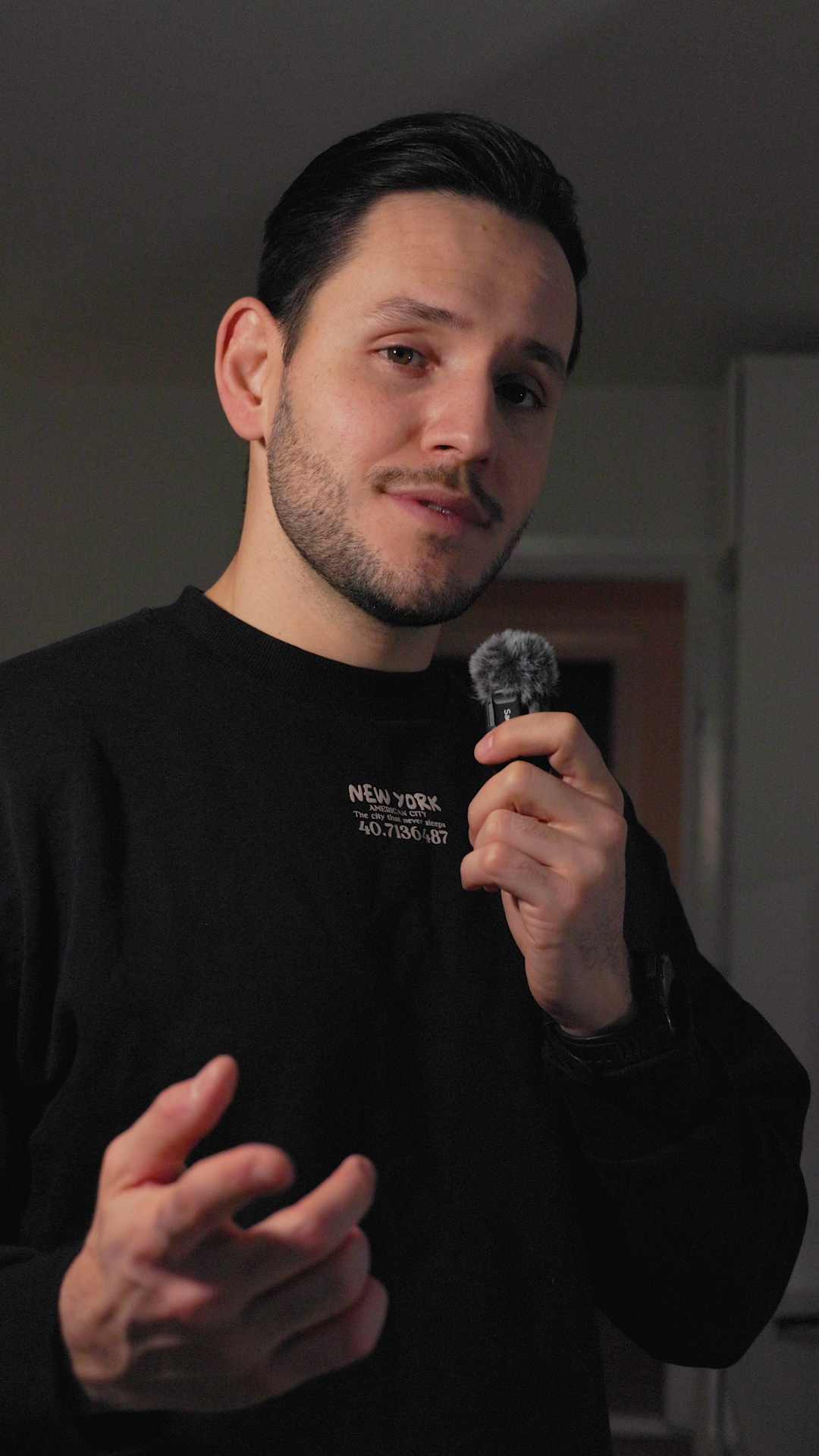 A young man with dark hair and facial hair holding a microphone while looking at the camera indoors.