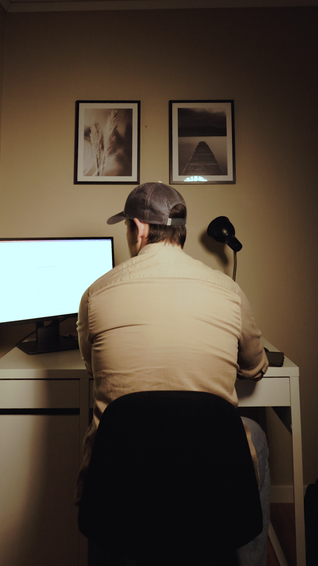 Man sitting at a desk with a computer monitor, facing away, wearing a cap and beige shirt, in a room with framed pictures of a palm tree and a pier on the wall.