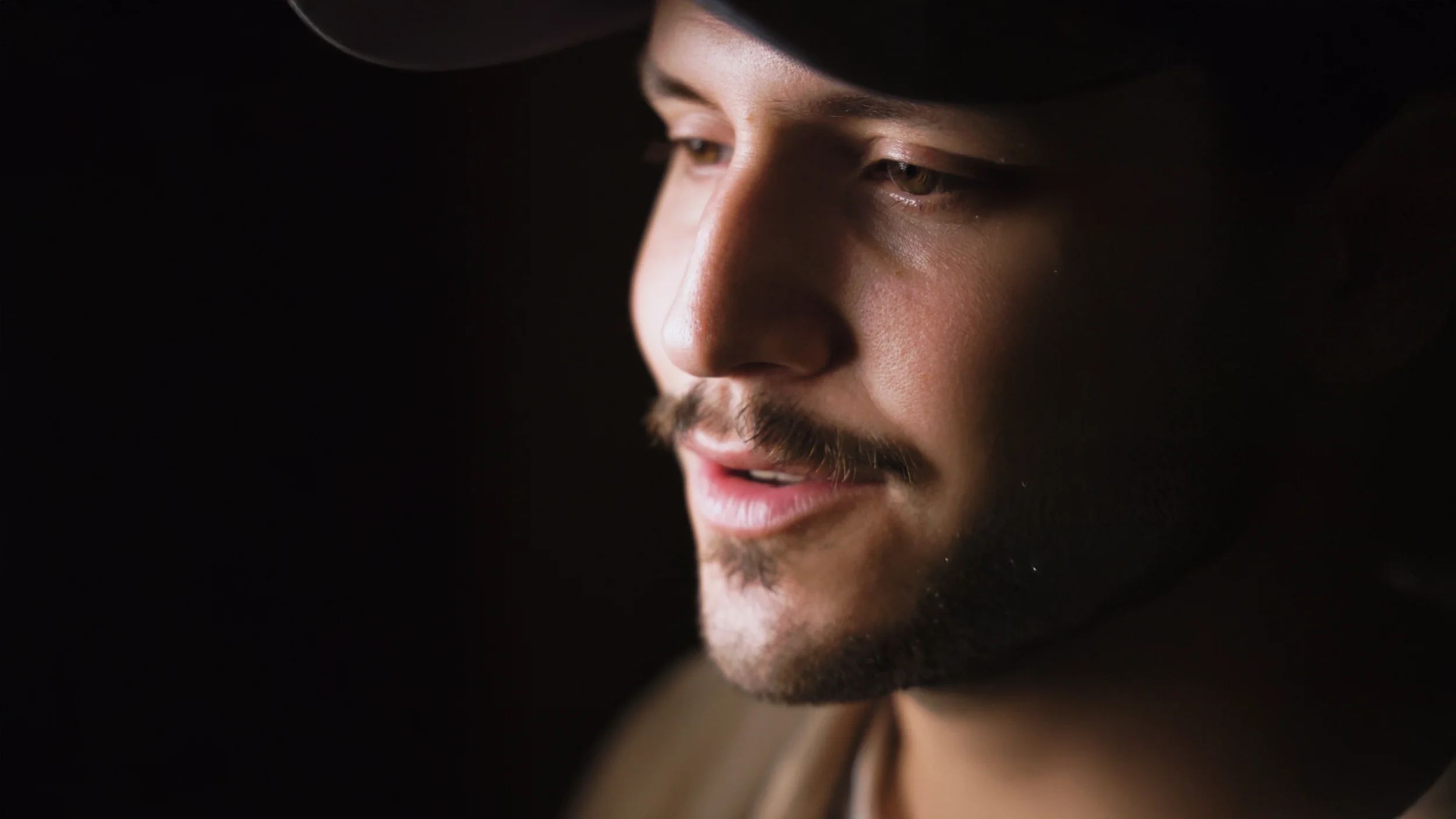 Close-up of a young man's face with a beard, mustache, and wearing a black hat, looking down thoughtfully against a dark background.