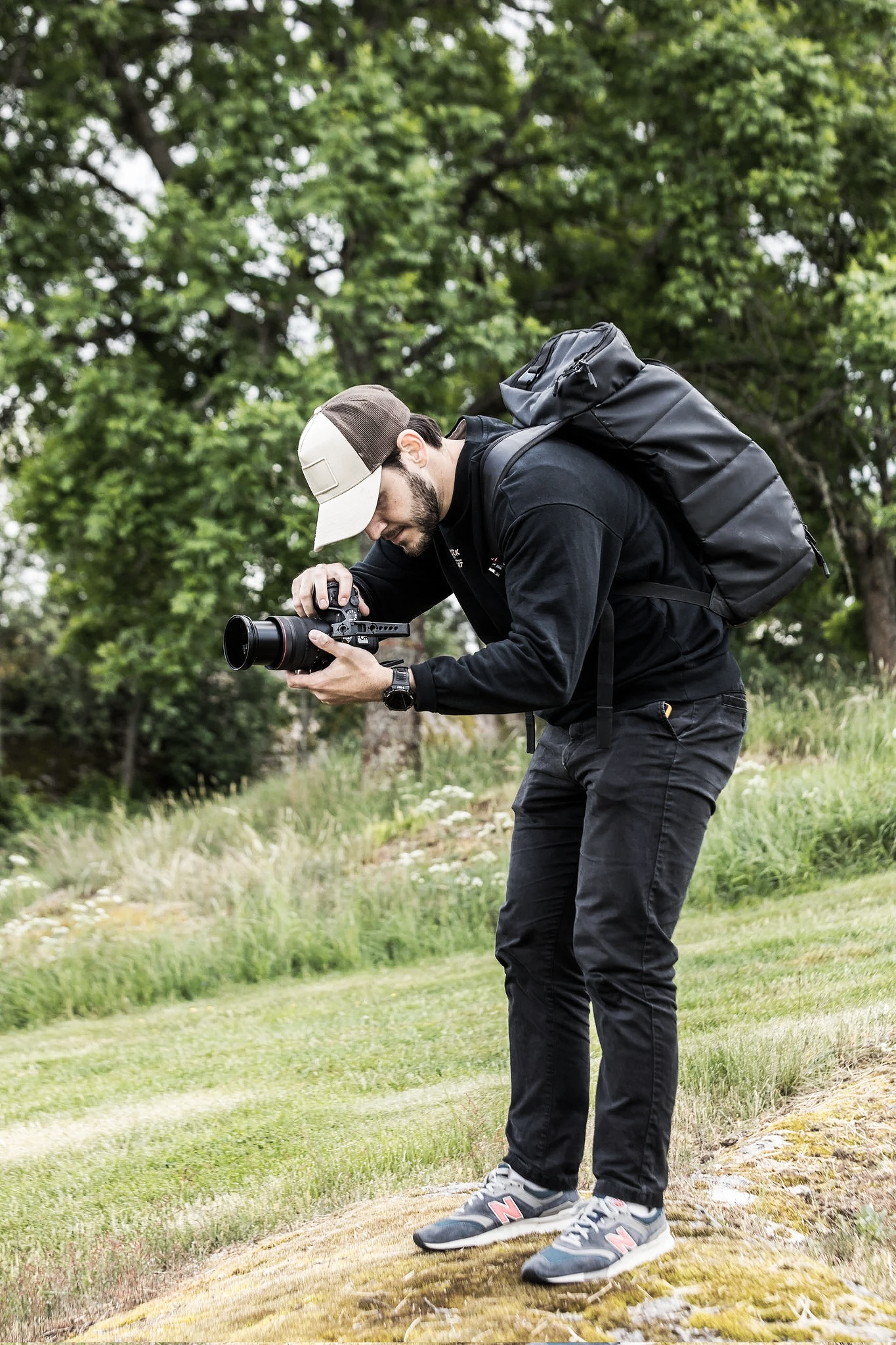 A man outdoors on a grassy area, taking pictures with a professional camera, wearing a black jacket, gray sneakers, a beige and dark brown cap, and carrying a large black backpack.