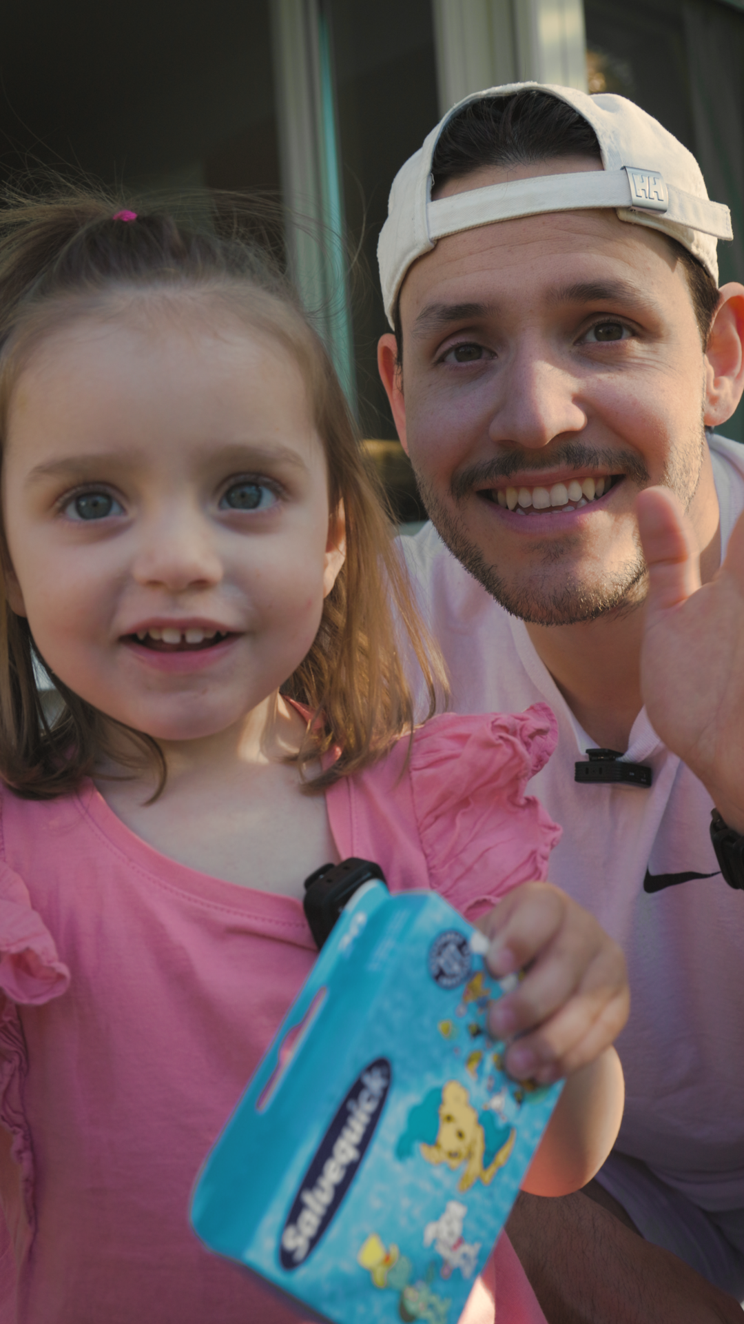 A young girl and a man, possibly her father, smiling and waving at the camera. The girl is holding a blue snack pack featuring cartoon characters.