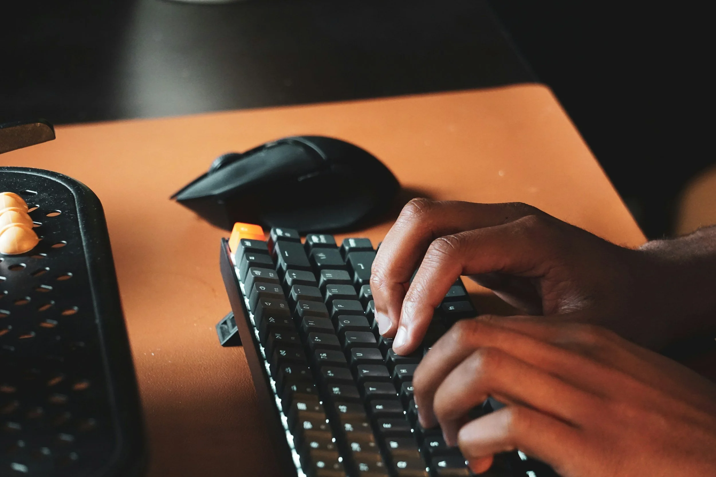 A person's hand typing on a black computer keyboard on a wooden desk with a wireless mouse in the background.