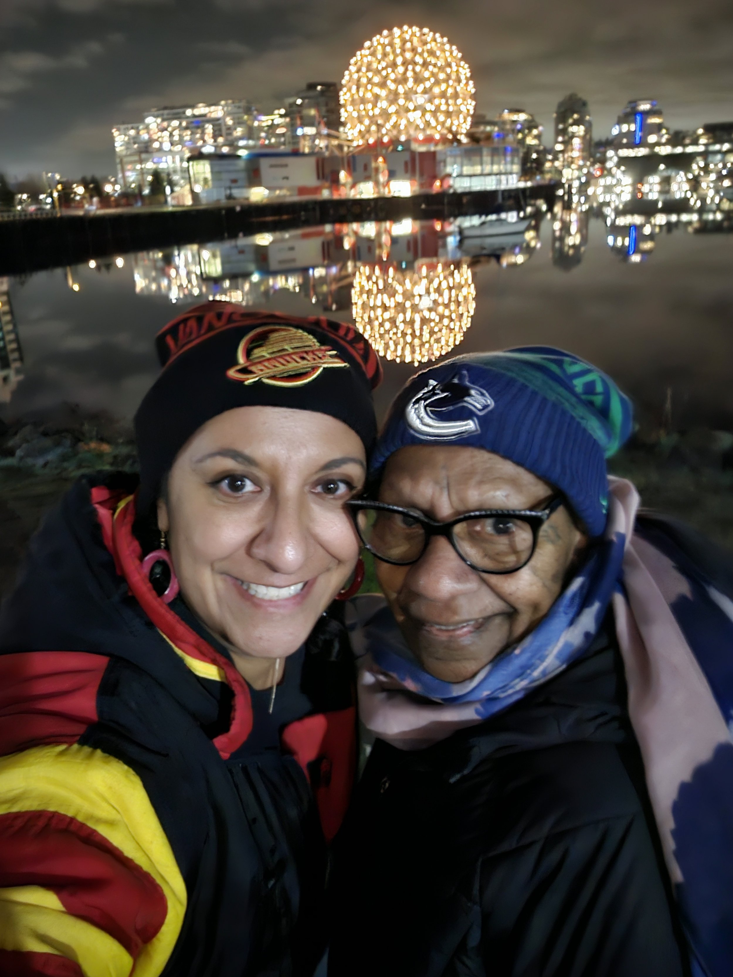 Two smiling women taking a selfie with a cityscape and illuminated spherical structure reflected in a body of water at night in the background.