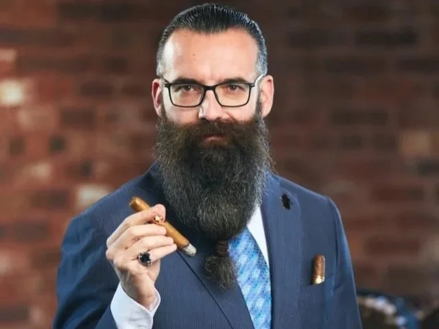 Ein Mann mit Brille, Vollbart und Anzug, der eine Zigarre hält, vor einem Ziegelsteinwand-Hintergrund.