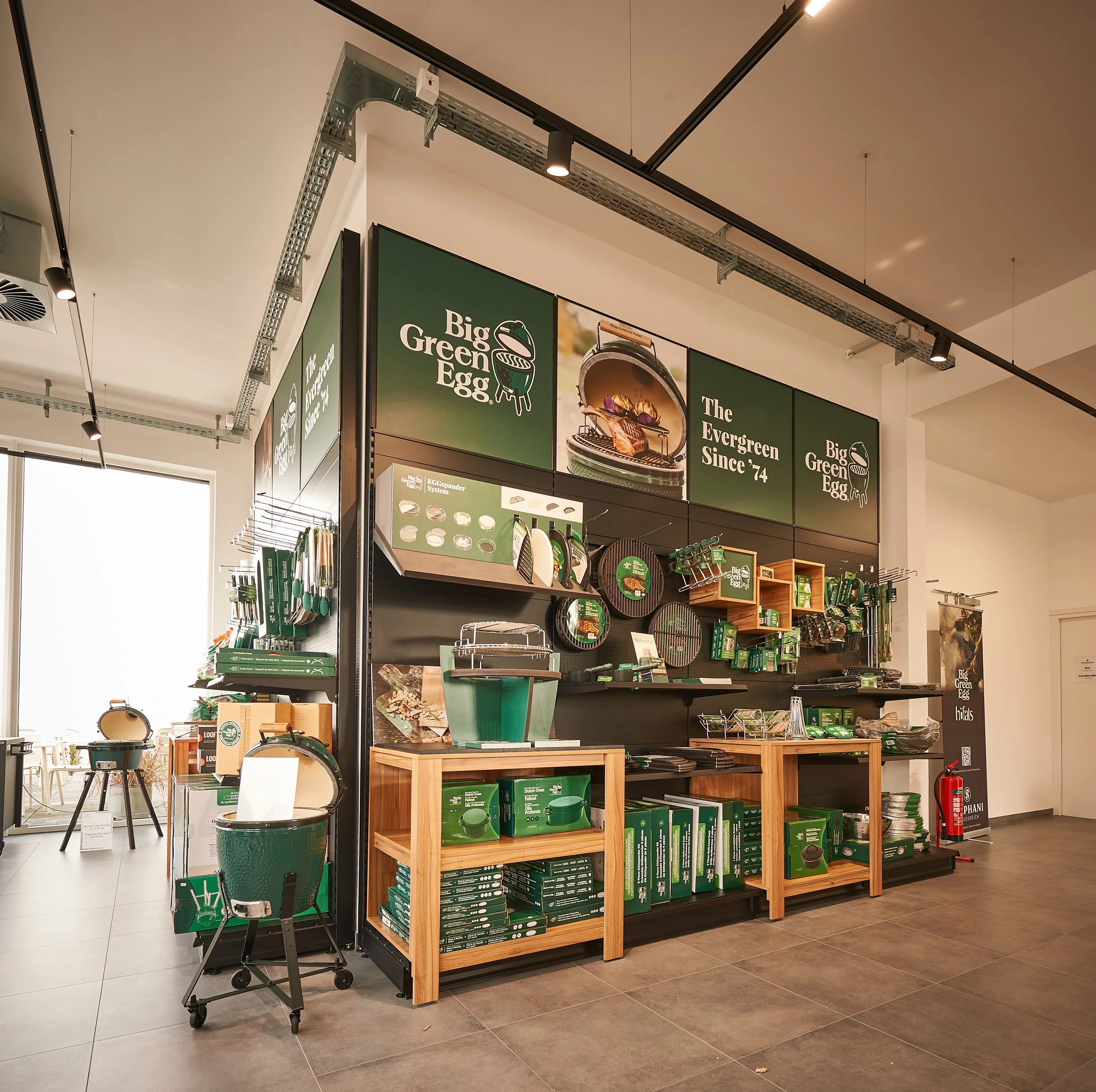 Eine Verkaufswand mit vielfältigem Gartenzubehör und Grillutensilien, überwiegend in grüner Farbe, in einem Geschäft. Wände sind mit Plakaten, die die Marke "Big Green Egg" und die Geschichte des Unternehmens zeigen, dekoriert.