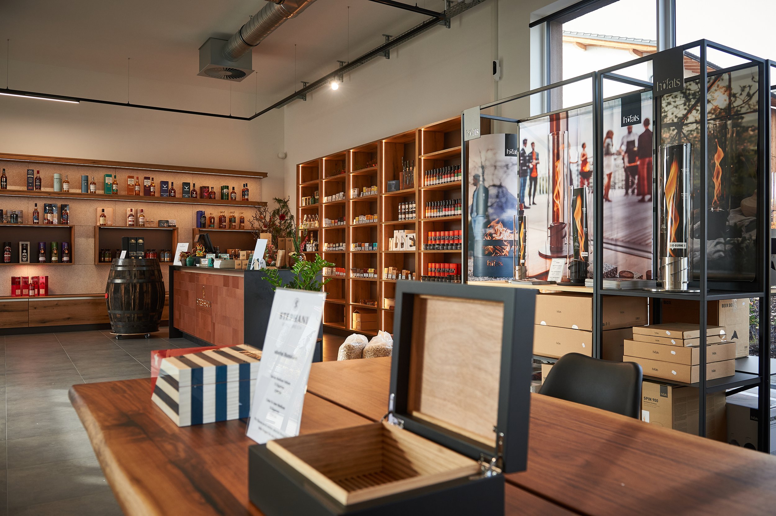 Innenraum eines Fachgeschäfts mit Holzwandregalen, Schrank mit Flaschen, Theke mit Kasse, Kartons und Verkaufsdisplays für Rauchwaren, große Fenster lassen Tageslicht herein.