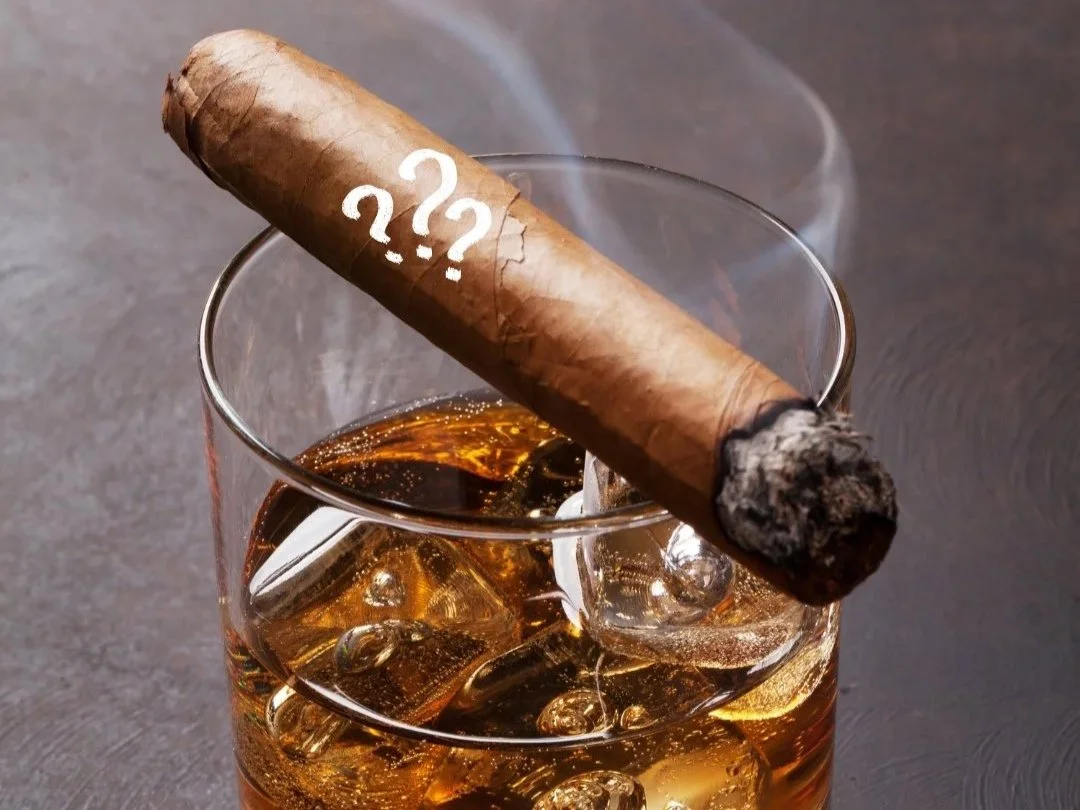 Ein Glas mit Whisky und Eiswürfeln, darauf eine angeschlagene Zigarre mit Asche und glimmender Spitze.