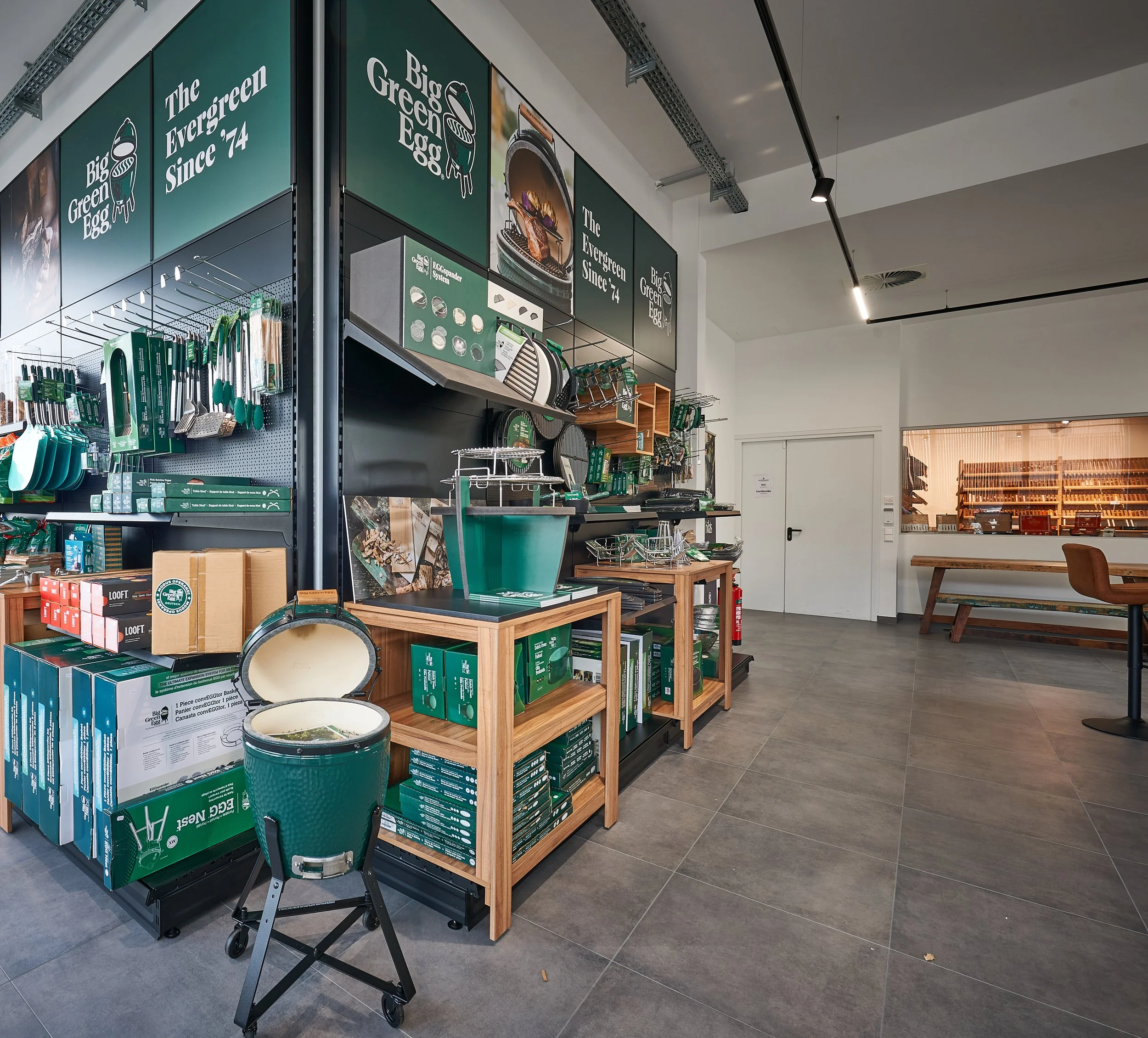 Ein Laden mit Gartengeräten, Werkzeugen und Zubehör, vor allem in Grün. Regale und Tische präsentieren Produkte, in der Nähe steht ein grüner Gartentopf. Im Hintergrund ist ein Raum mit viel Holzreinigung.