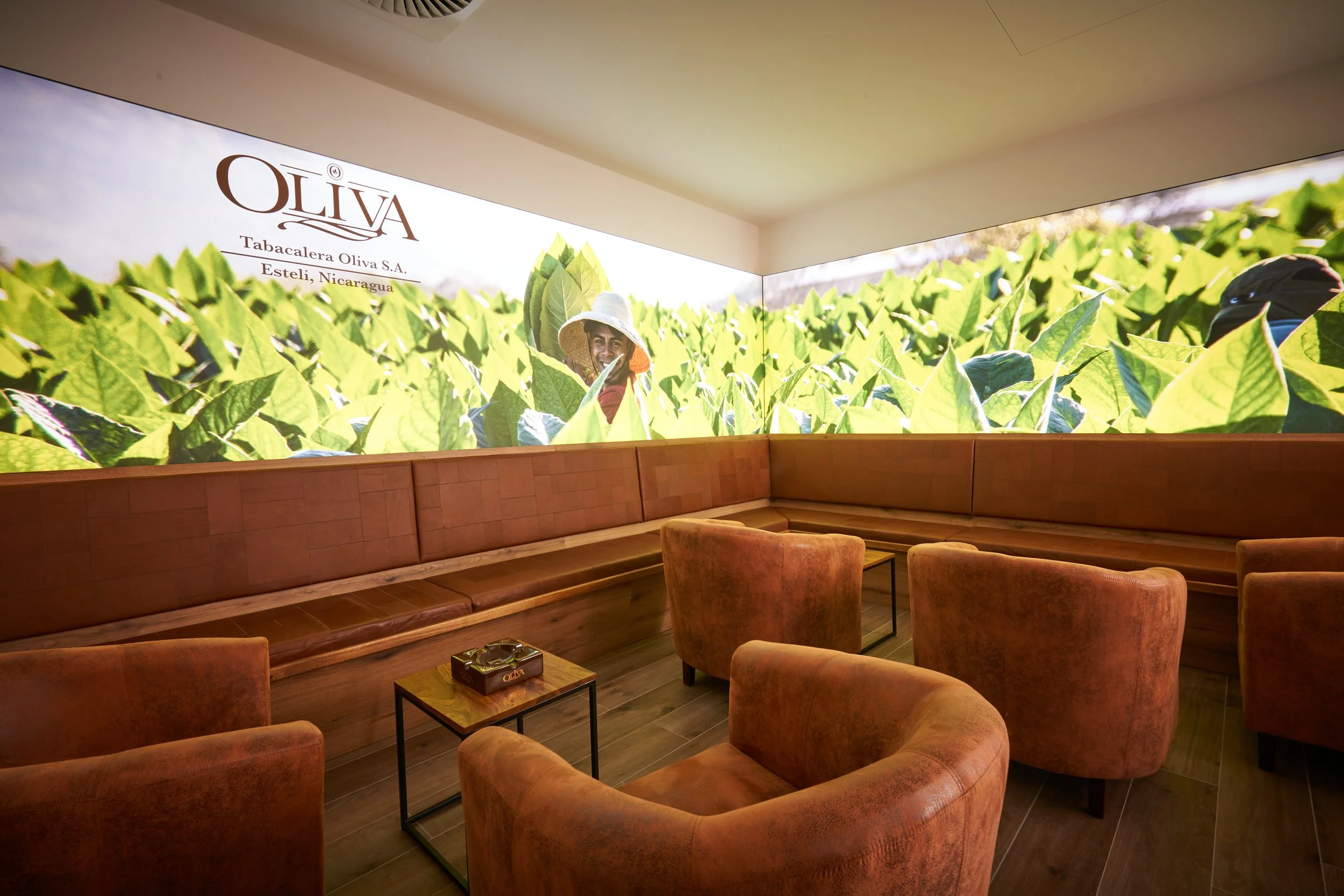 Ein Lounge-Bereich mit braunen Sesseln und einer langen braunen Sitzbank vor einer Wand mit einem großen Wandbild, das eine Kakaopflegeplantage zeigt, darunter steht 'OLIVA Tabacalera Oliva S.A. Estelí, Nicaragua'.