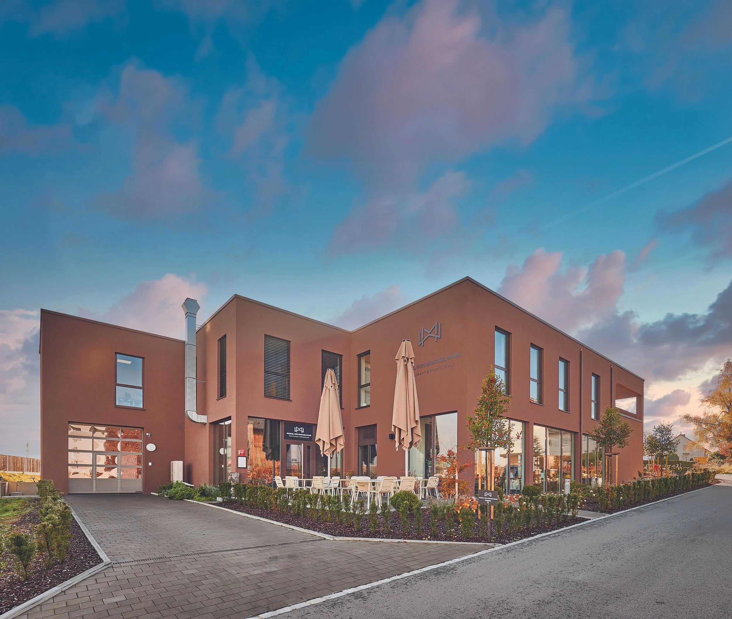 Modernes braunes Gebäude mit großen Fenstern, Sitzgelegenheiten im Freien und begrüntem Gartenbereich bei Sonnenuntergang.